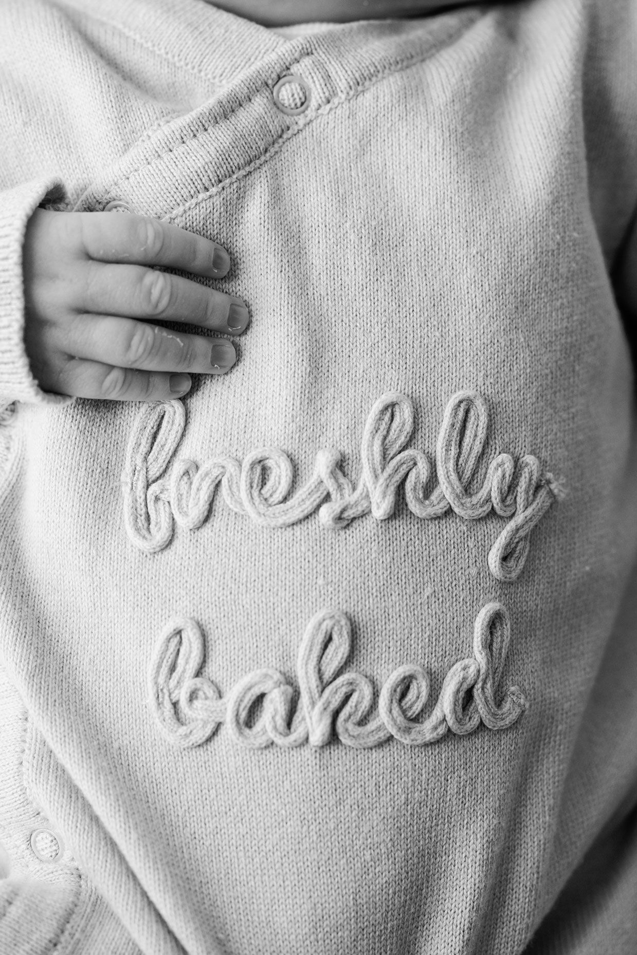Black and white close-up of a newborn's tiny hand resting on a knit sweater embroidered with the words freshly baked.