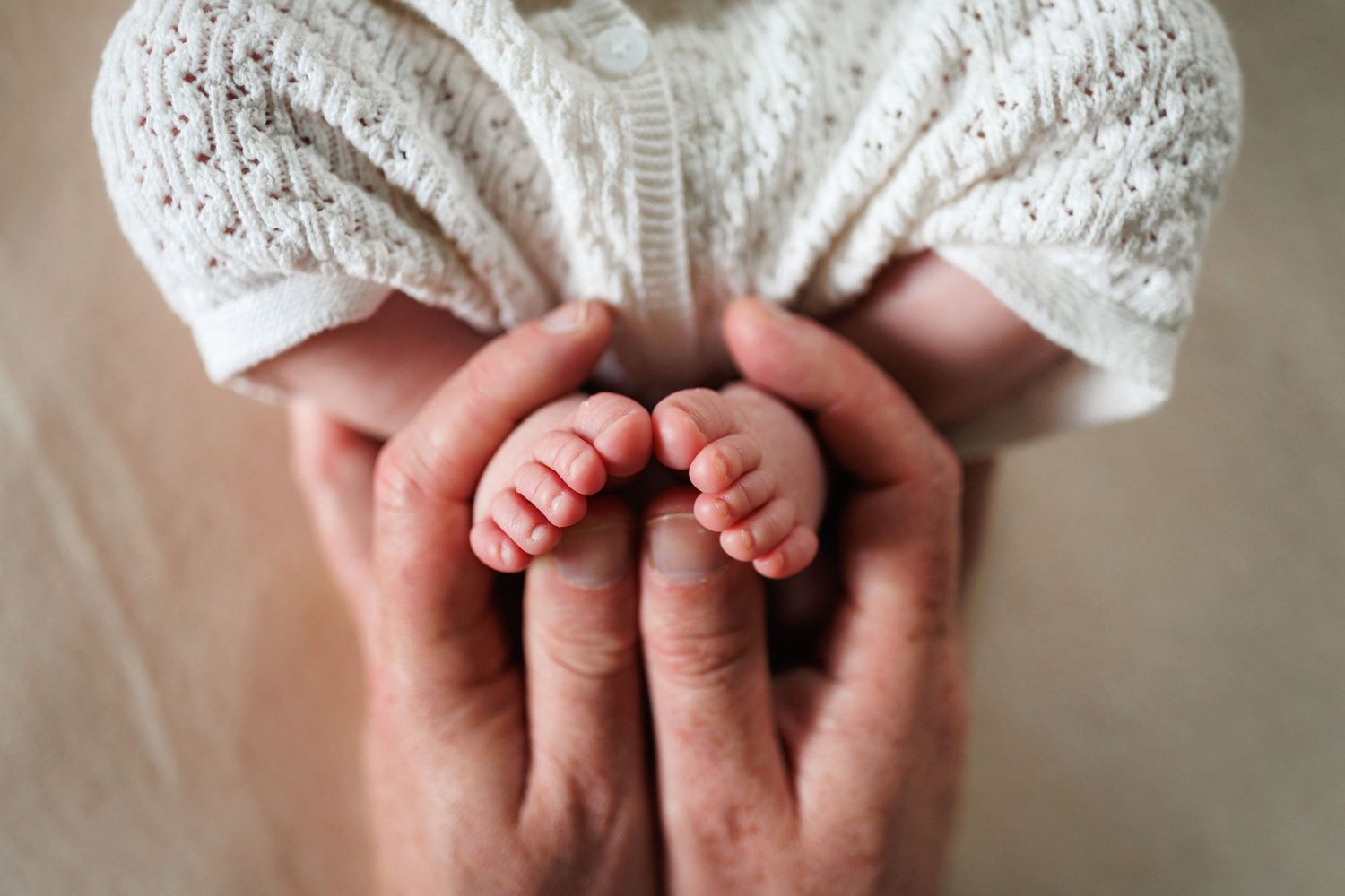 Close-up of parent hands tenderly holding tiny newborn baby feet in white knit romper.