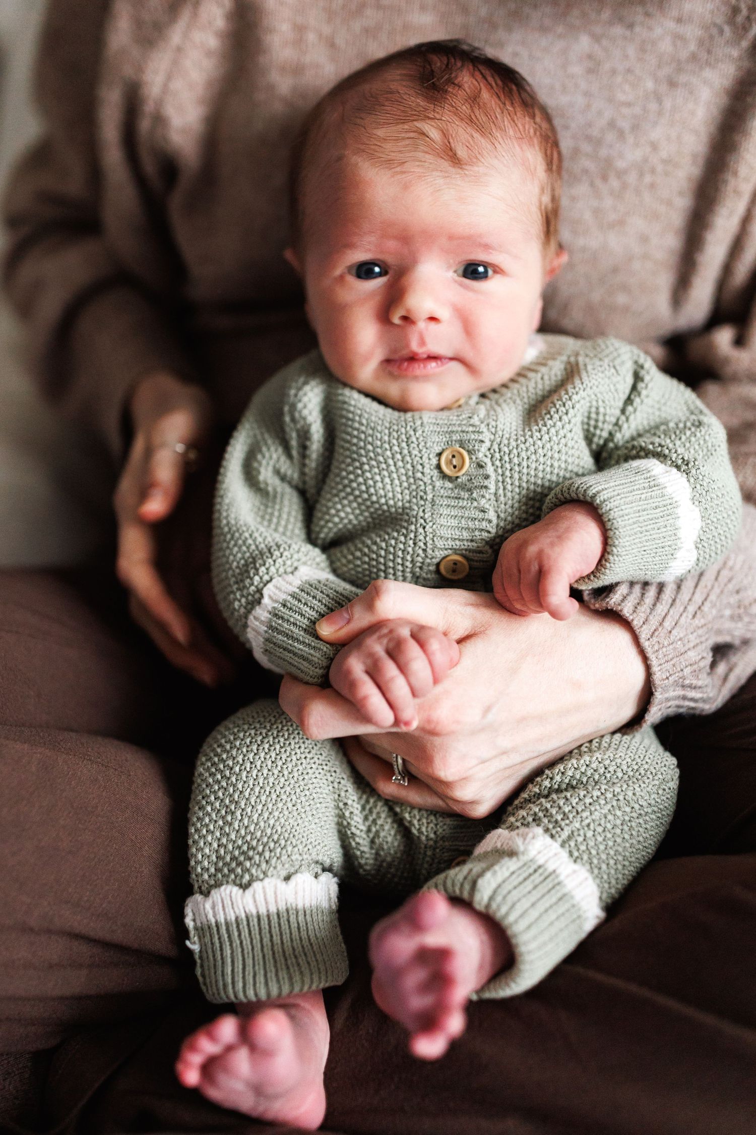 Alert newborn baby in a sage green knit romper sits supported on a parents lap looking at the camera.