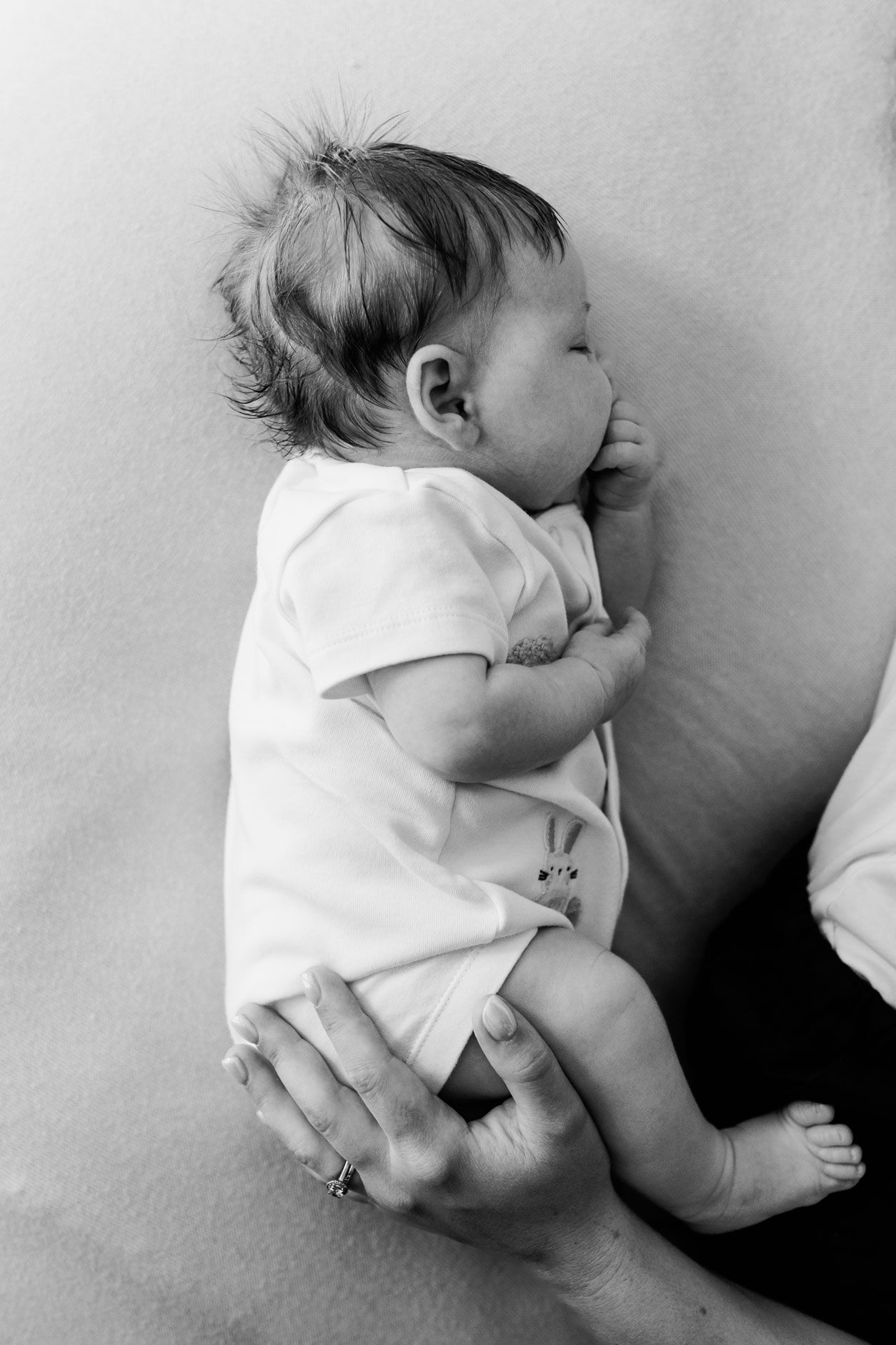 Black and white close-up of a sleeping newborn baby in a white onesie being held gently.