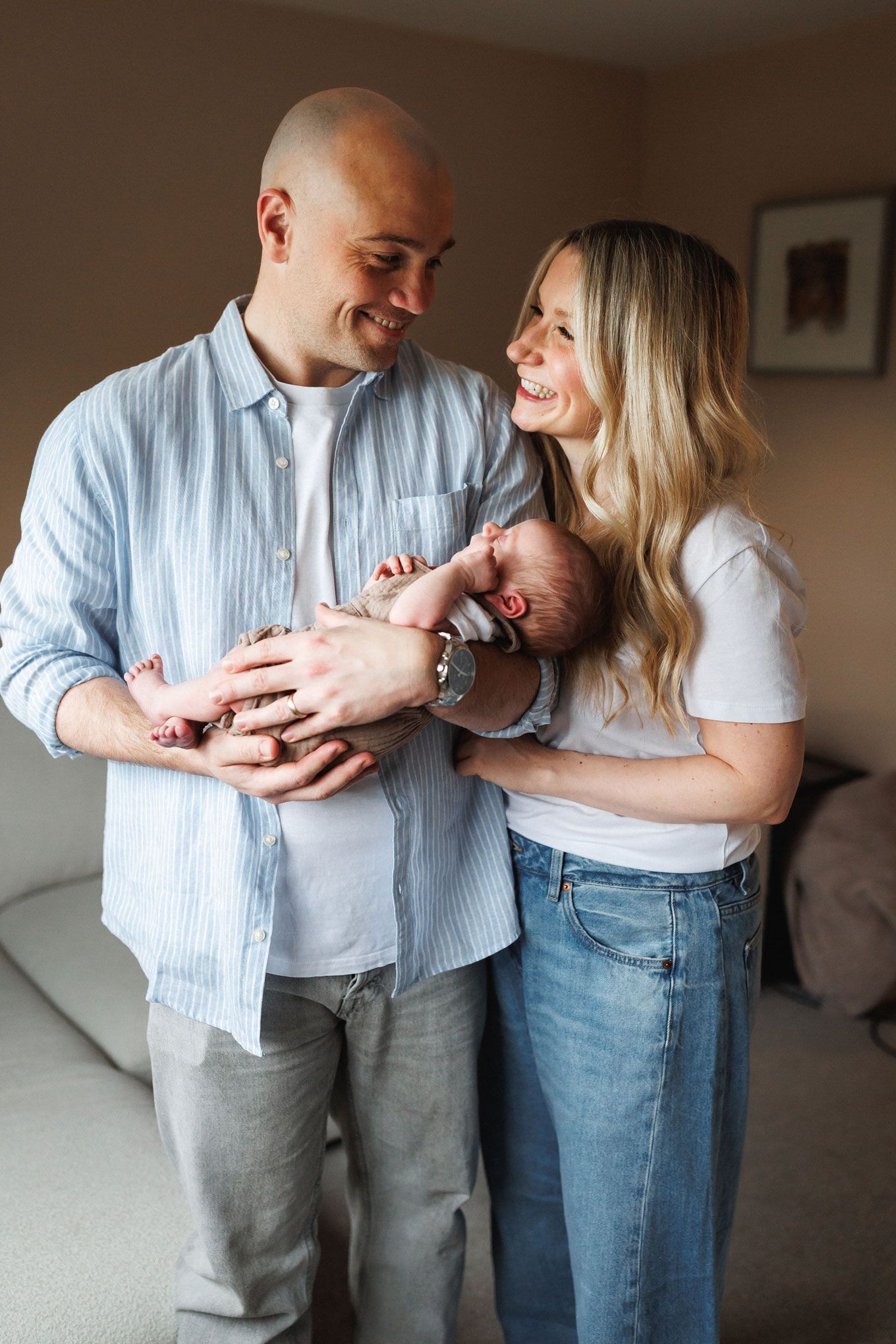 A smiling couple stands indoors holding their newborn baby, with the bald man and blonde woman gazing at each other lovingly.