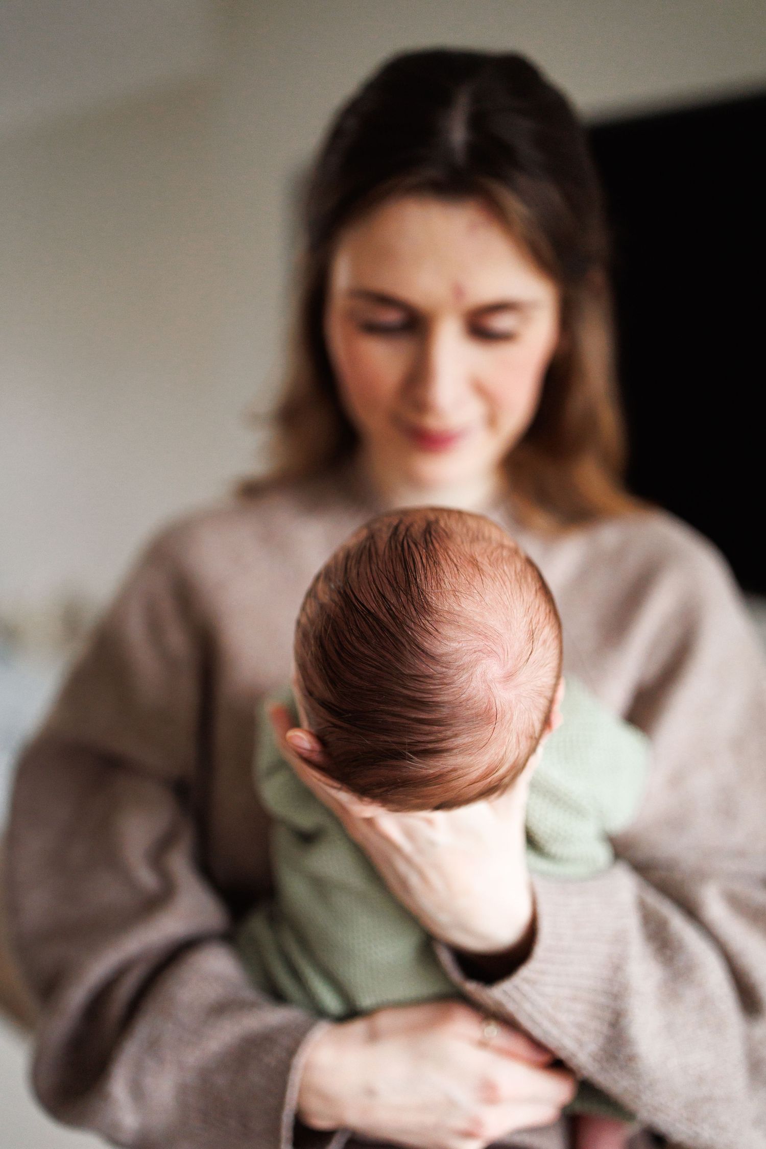 A mother in a beige sweater tenderly holds her newborn baby facing away from the camera indoors.
