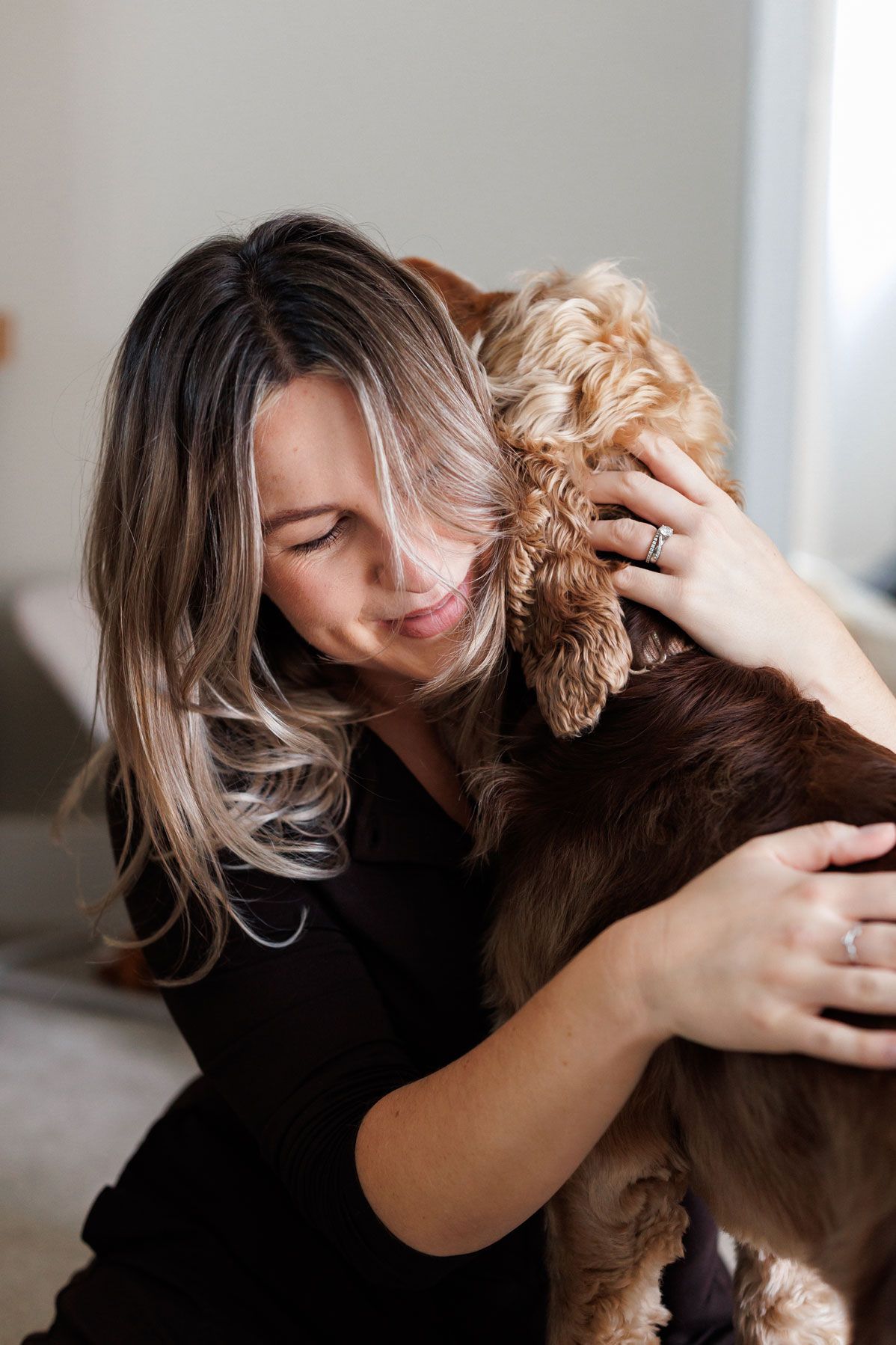 Woman with long highlighted hair closes her eyes and lovingly hugs a brown and tan cocker spaniel in a bright, cozy room.