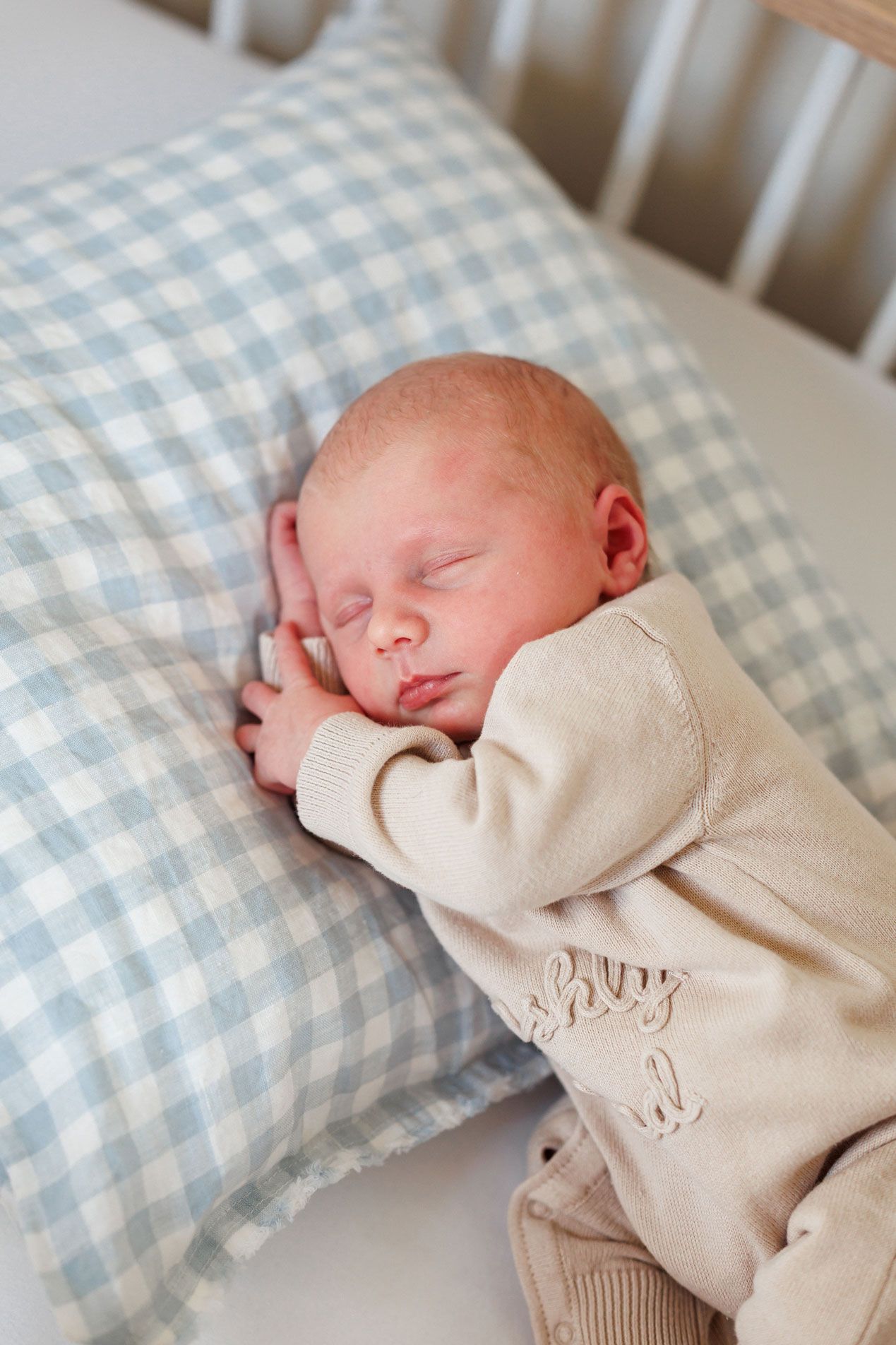 Sleeping newborn baby in a cream knit outfit resting on a blue and white gingham pillow in a white crib.