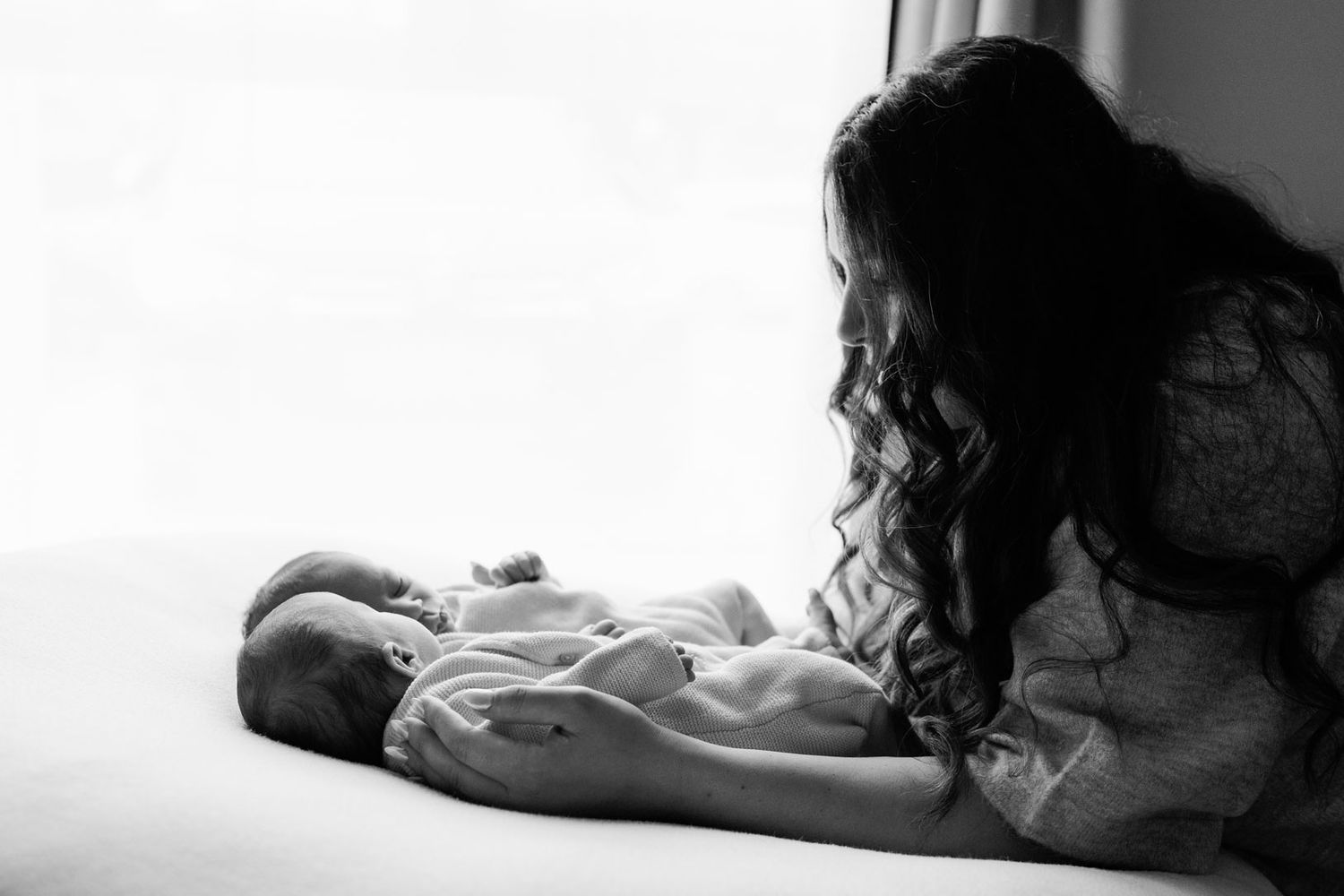 Black and white photo of a long-haired mother gazing lovingly at her sleeping newborn twins lying side by side on a bed by a bright window.