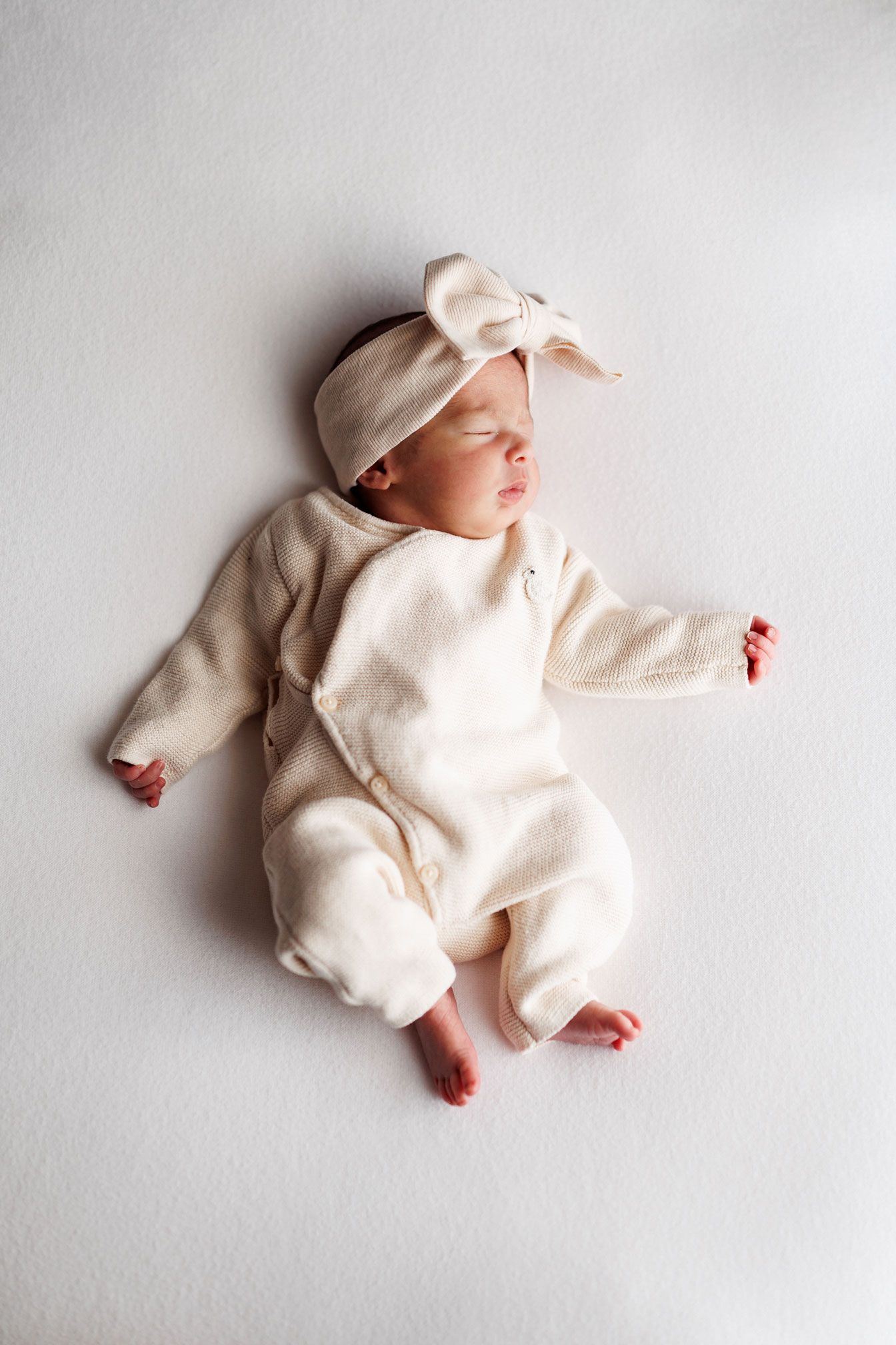 Sleeping newborn baby in a cream knit wrap romper and matching bow headband lying on a soft white background.