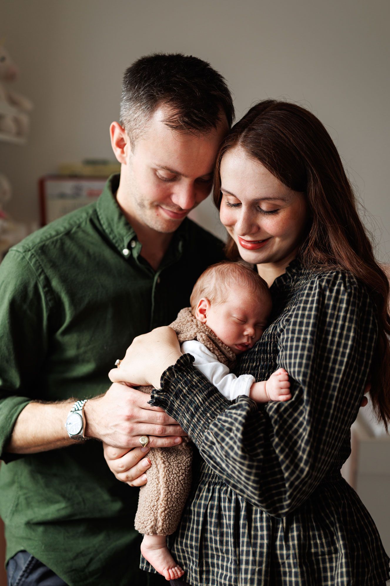 Smiling parents in green and plaid gently cradle their sleeping newborn baby in a cozy, softly lit nursery.