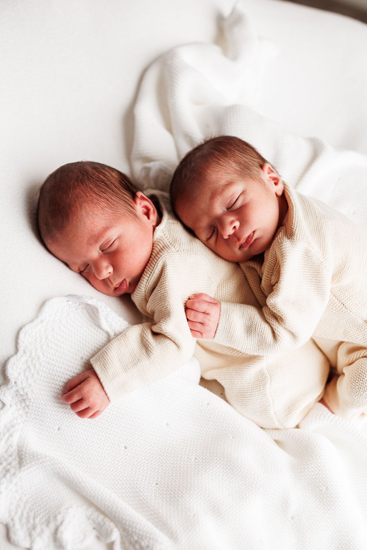 Two sleeping newborn babies in cream knit outfits lying side by side on a white blanket.