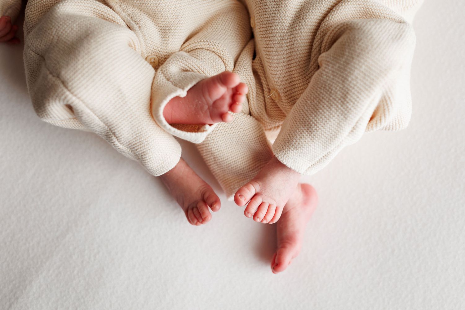 Close-up of a newborn baby's tiny bare feet and toes crossed in a cream knit outfit on a soft white blanket.