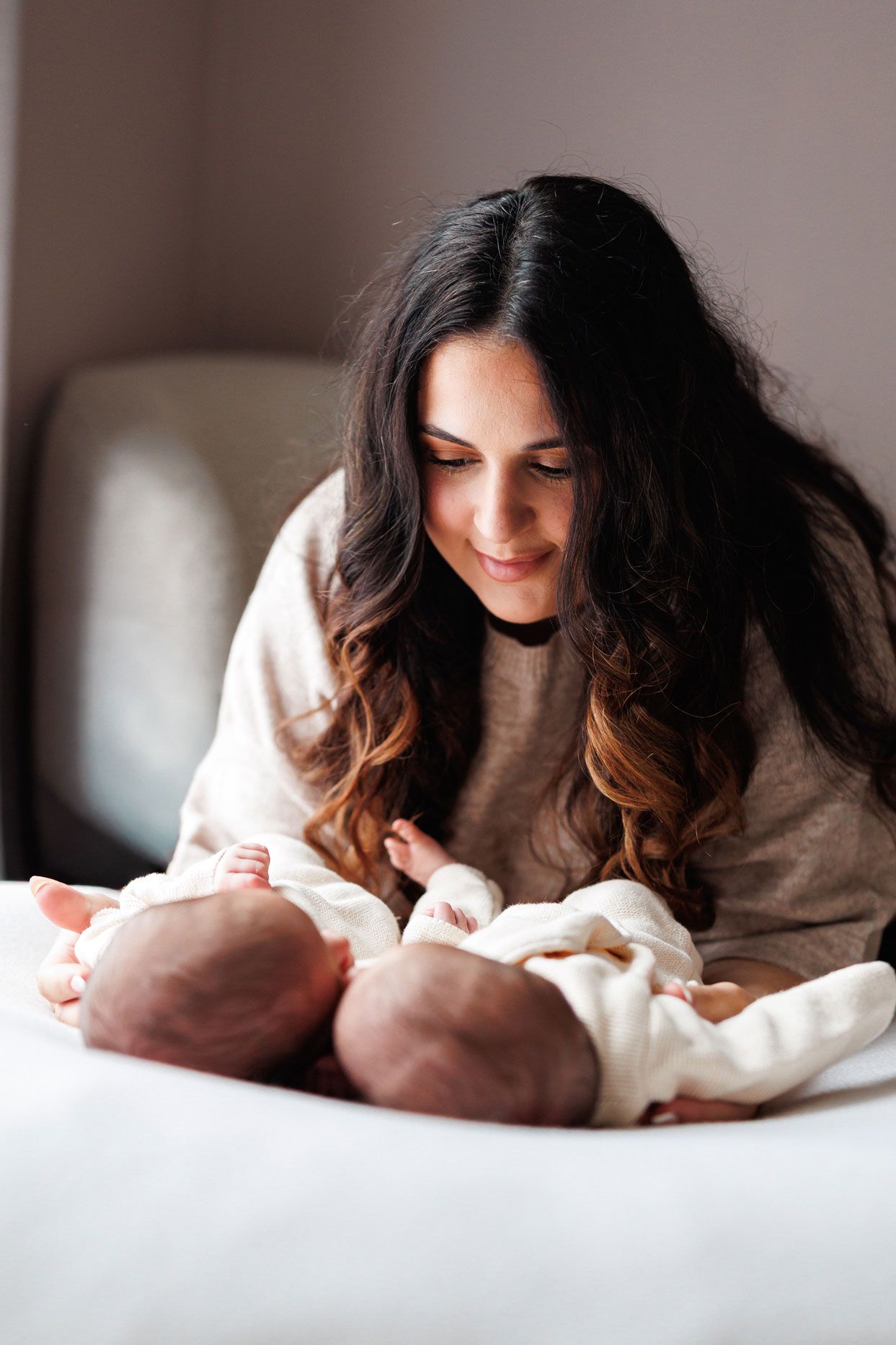 Smiling mother with long dark wavy hair gazes lovingly at two newborn twin babies lying side by side on a white bed.