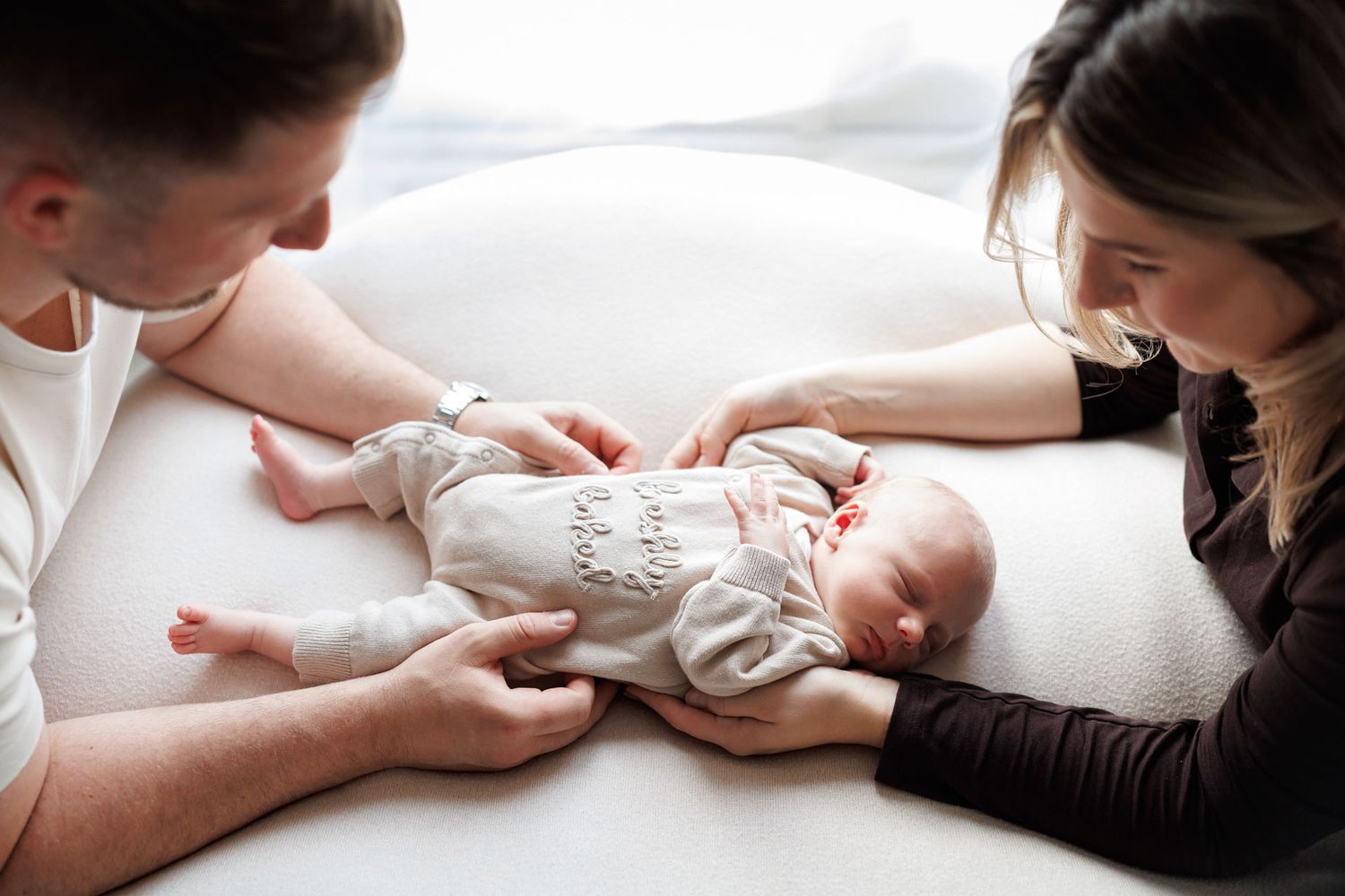 Parents gently hold a sleeping newborn baby in a cream knit outfit lying on a soft white cushion during a cozy newborn photo session.