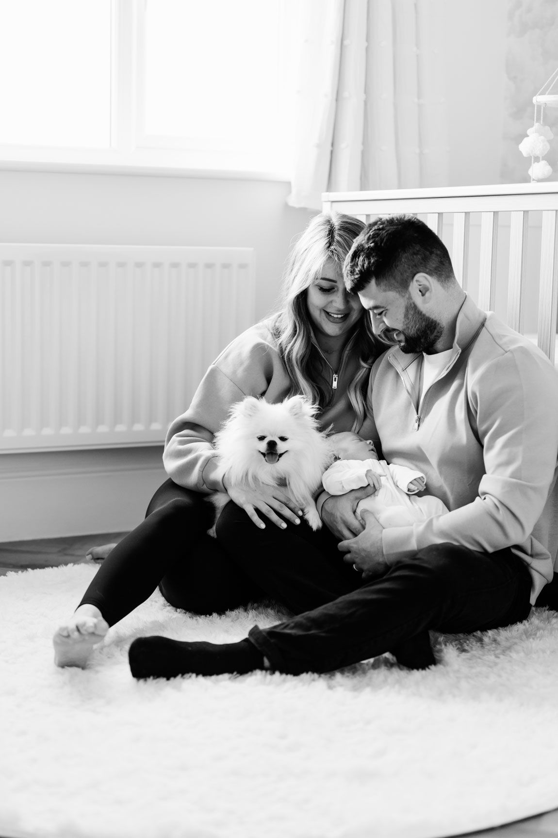 Black and white photo of a couple sitting on a fluffy rug with a white Pomeranian dog and newborn baby.