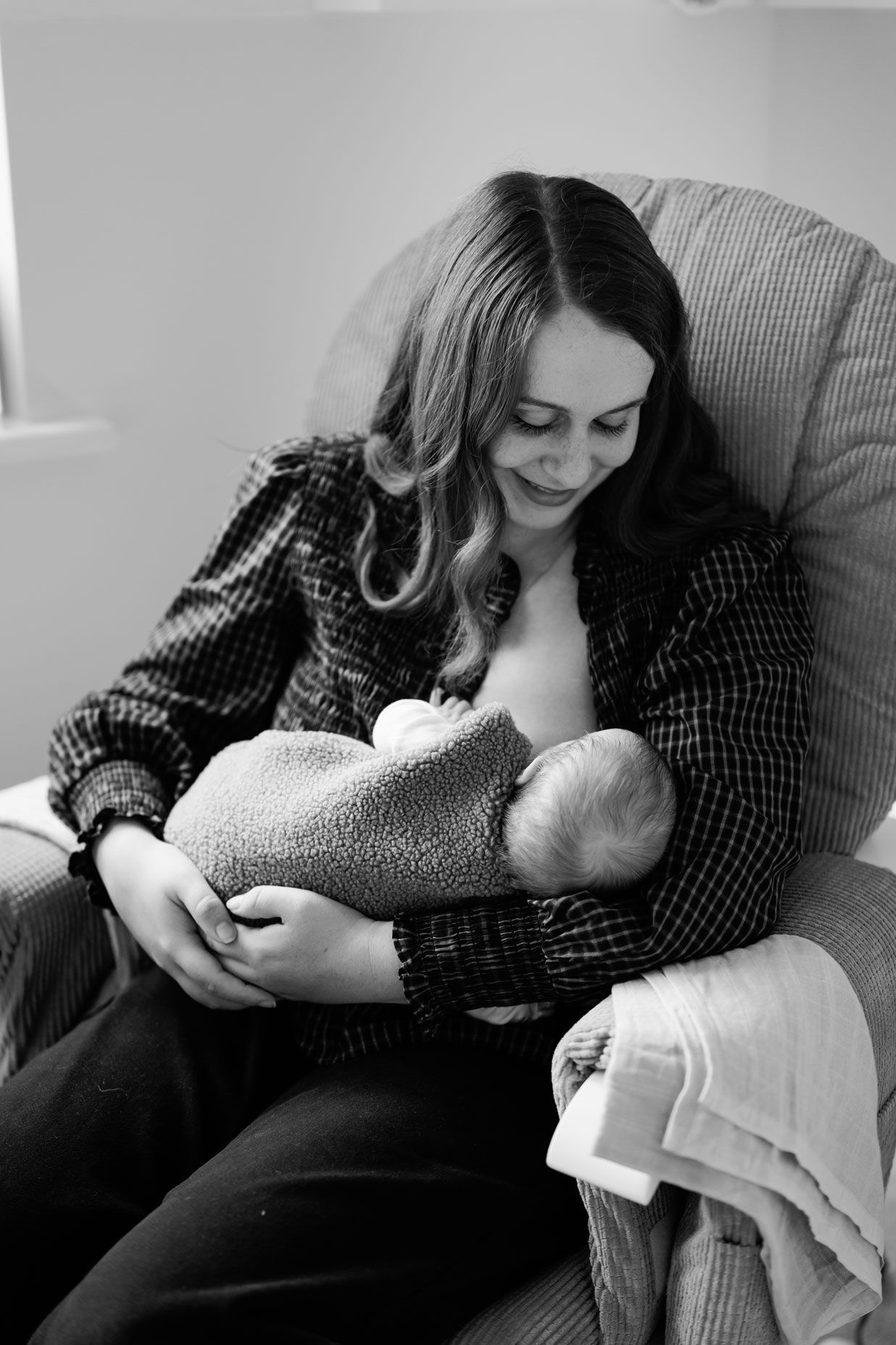 A smiling woman with long wavy hair sits in a cushioned armchair while nursing her newborn baby wrapped in a soft blanket.