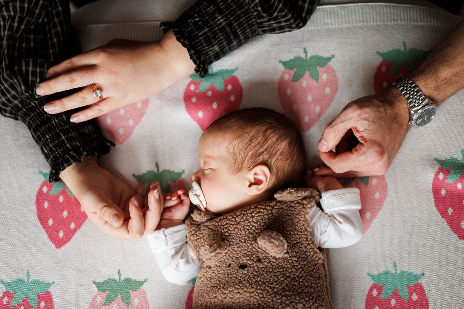 Sleeping newborn in a brown teddy bear outfit holds two adult hands on a soft blanket with a strawberry pattern.