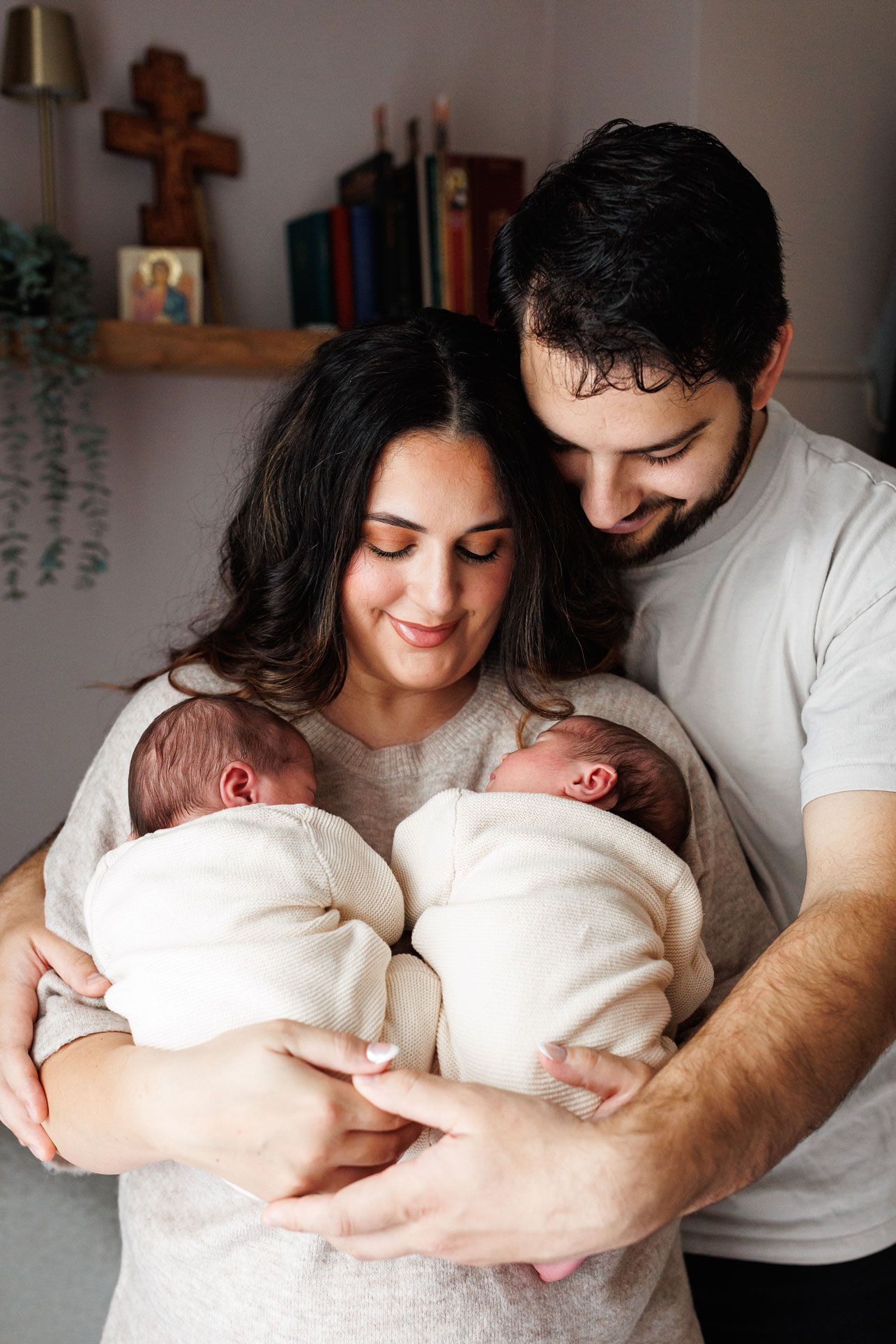 Smiling mother and father tenderly cradle two swaddled newborn twins in a cozy home with a wooden cross and books on a shelf.