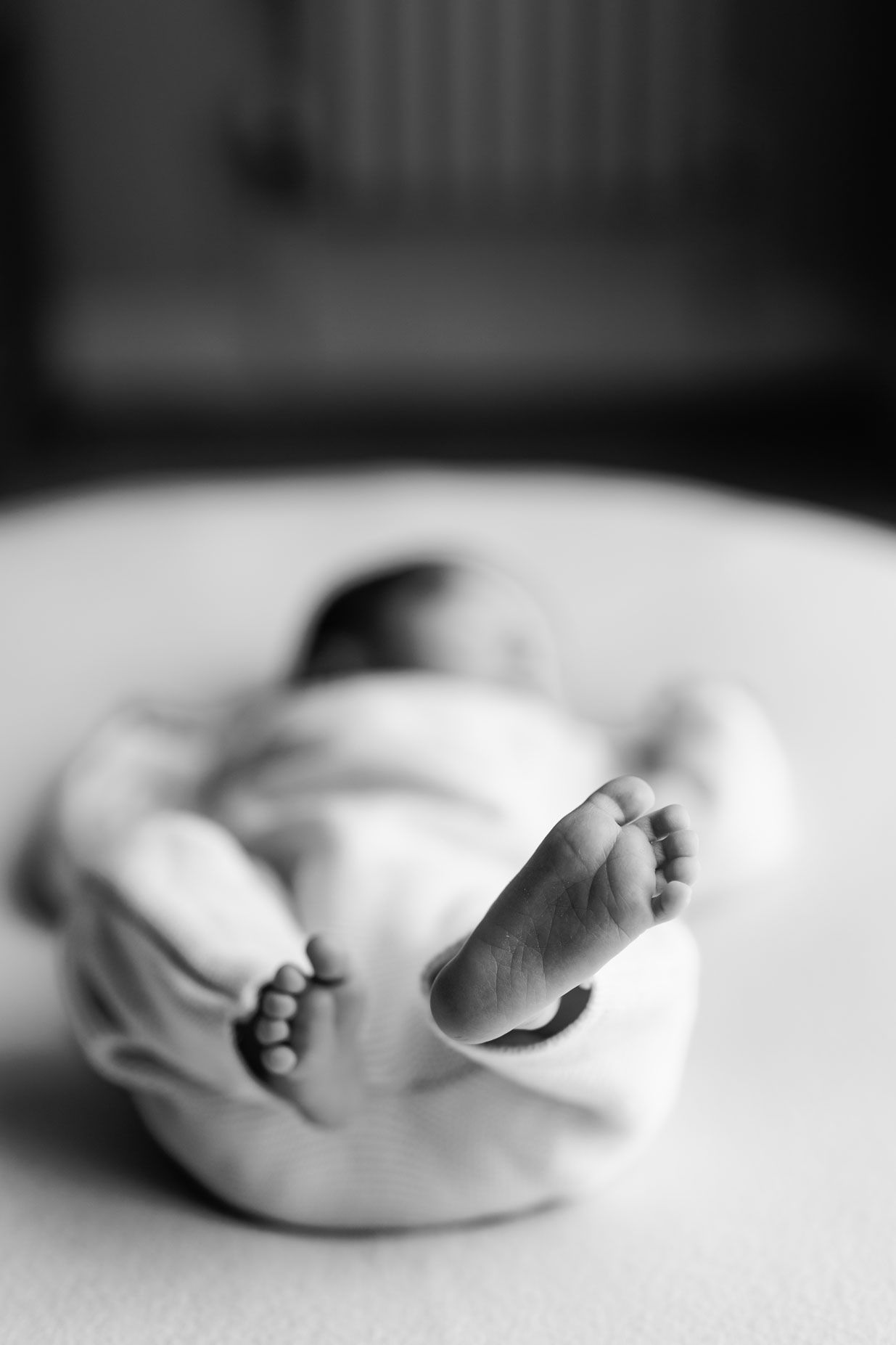 Black and white photo of a newborn baby lying on a soft blanket, with tiny feet in sharp focus and the swaddled body softly blurred.