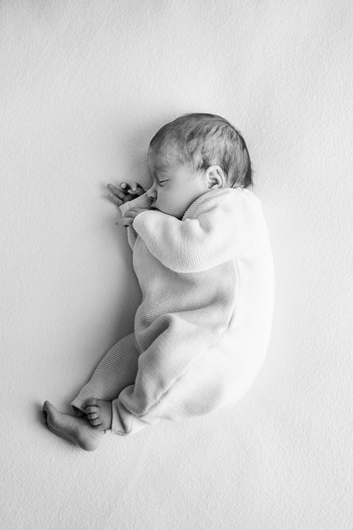 Black and white photo of a sleeping newborn baby in a knit outfit curled up on a soft blanket.
