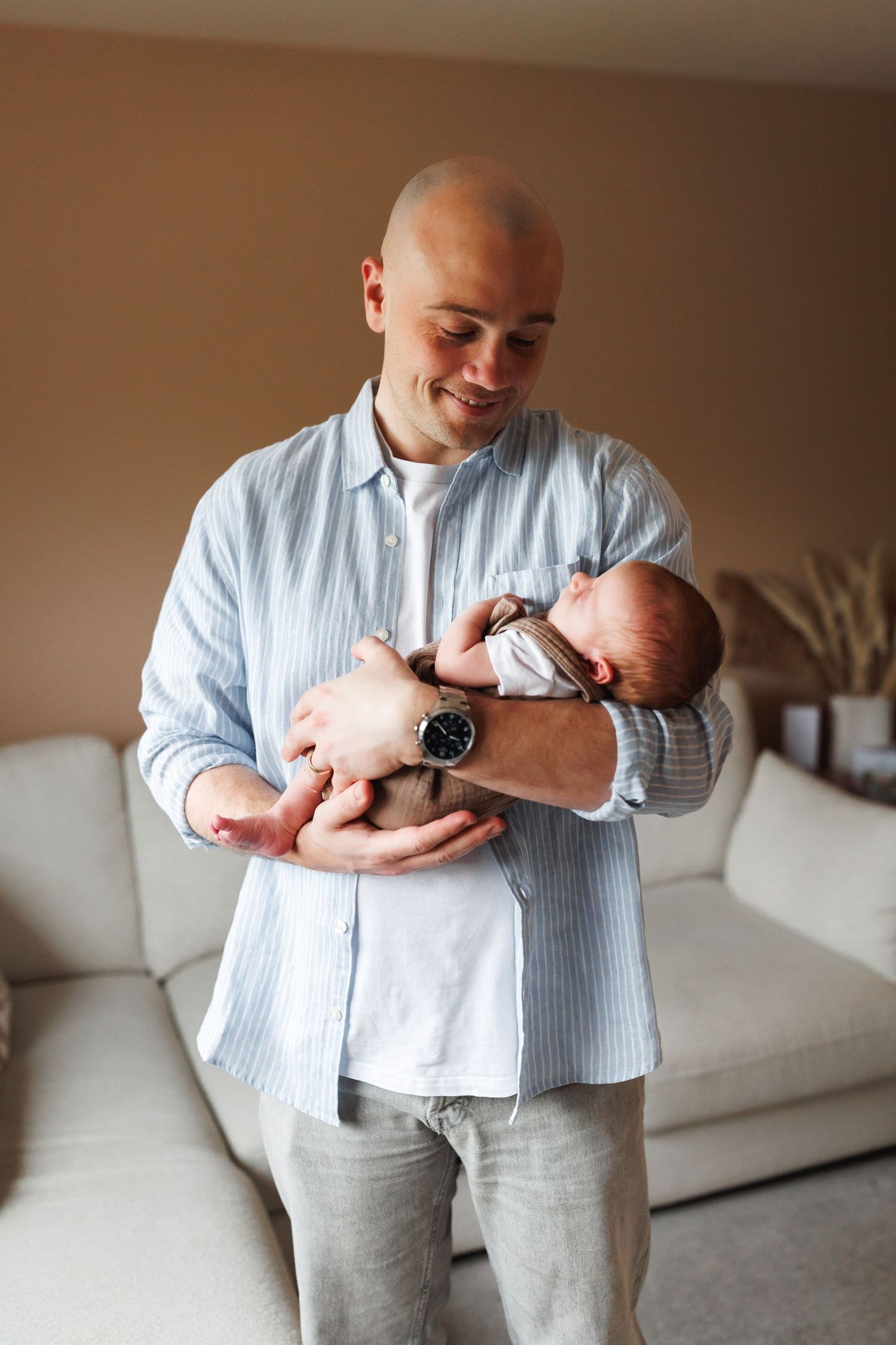 Proud father in blue striped shirt smiling while carefully holding his newborn baby at home.