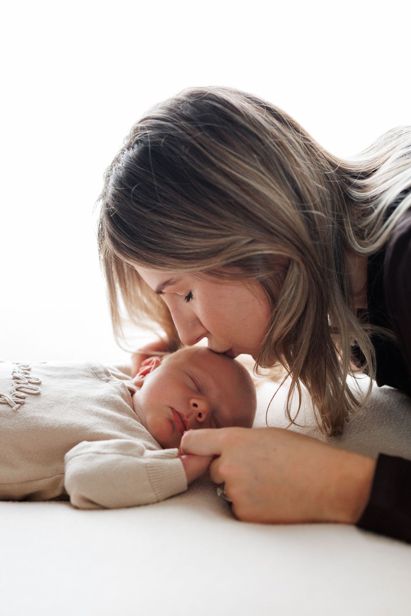 Mother with long blonde hair gently kisses the forehead of her sleeping newborn baby lying on a soft white blanket.