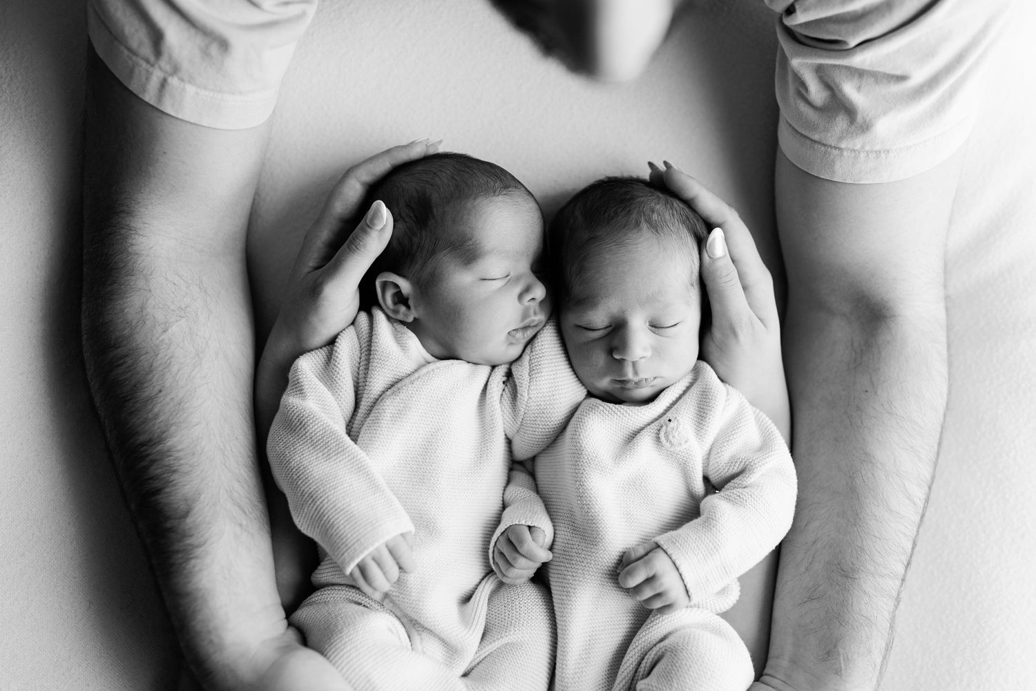 Black and white photo of two sleeping newborn twins in matching outfits cradled between a parent's hands and arms.