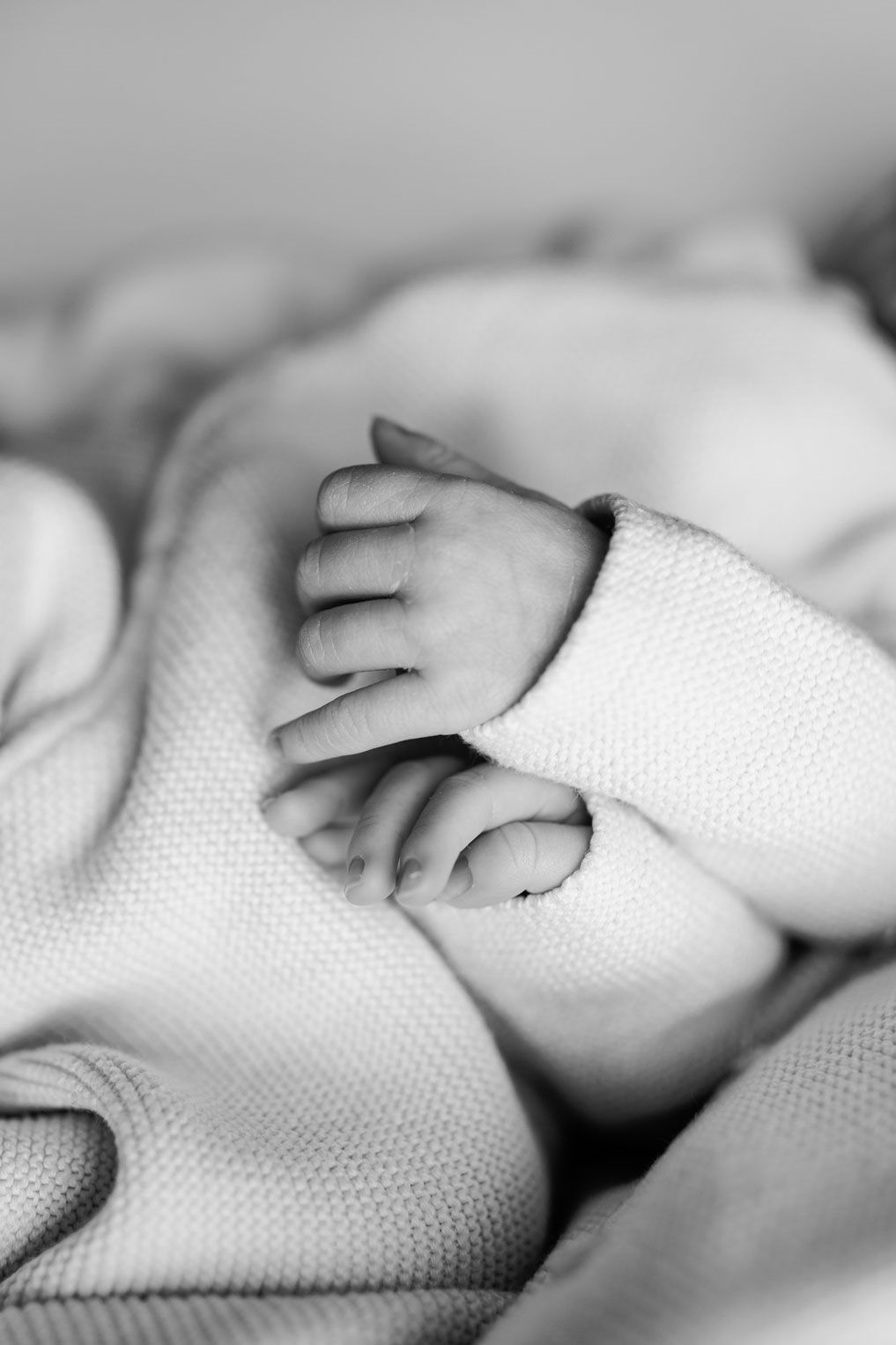 Black and white photo of a newborn baby's tiny hands peeking out from the sleeve of a soft knit sweater and blanket.