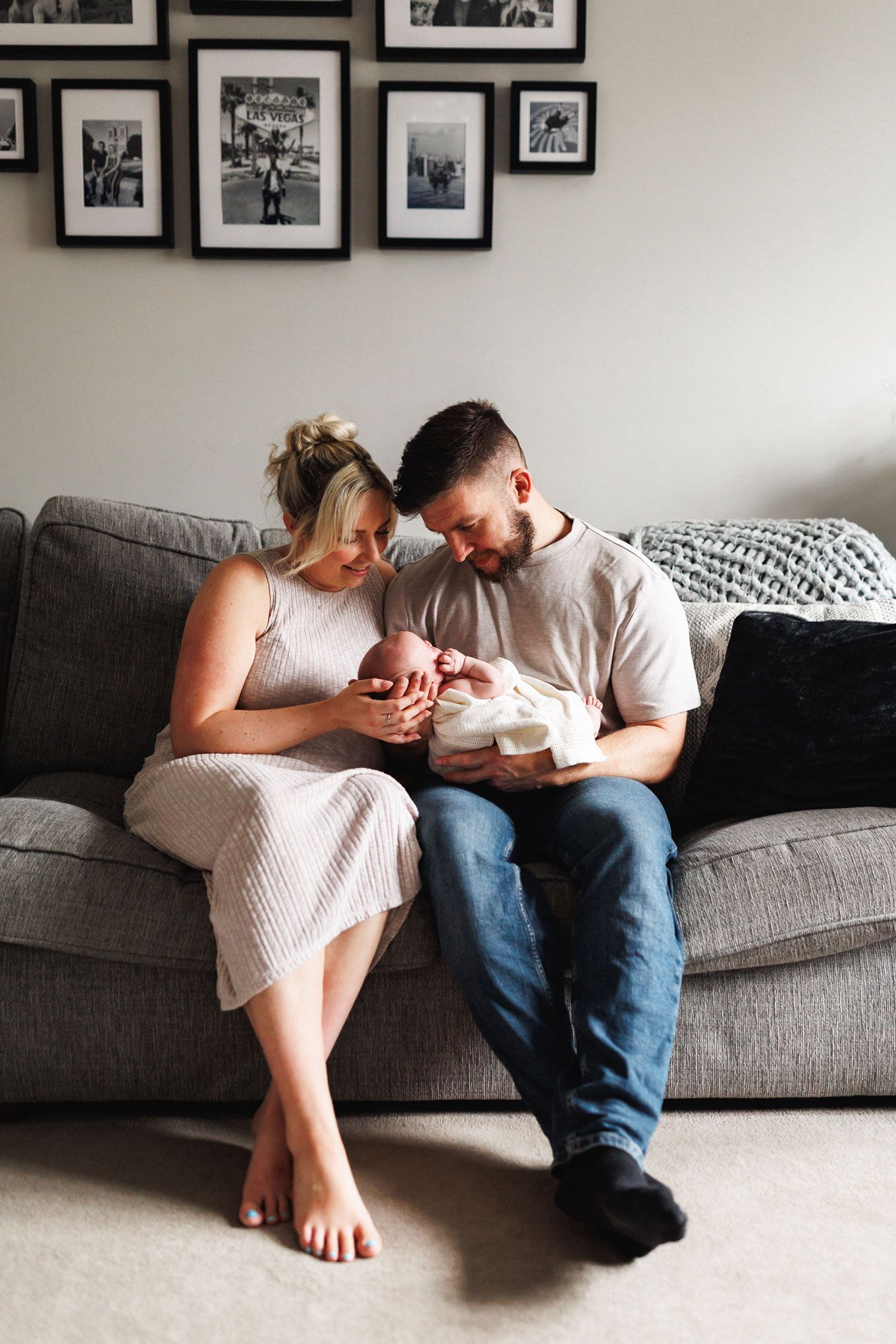 New parents sitting on a gray sofa, lovingly gazing at their newborn baby, with black and white family photos on the wall.