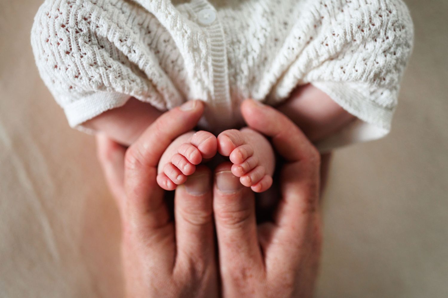 Close-up of adult hands gently cradling tiny newborn baby feet in a soft white knit outfit.