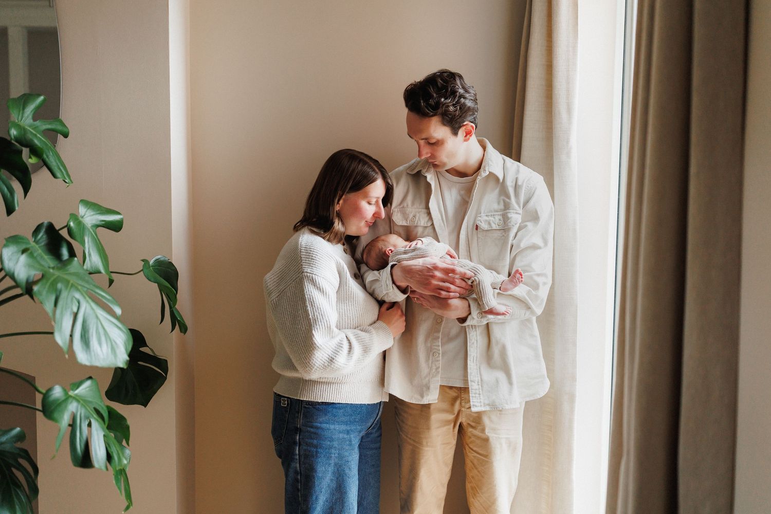 Young couple standing by window in soft light, lovingly gazing at their newborn baby.