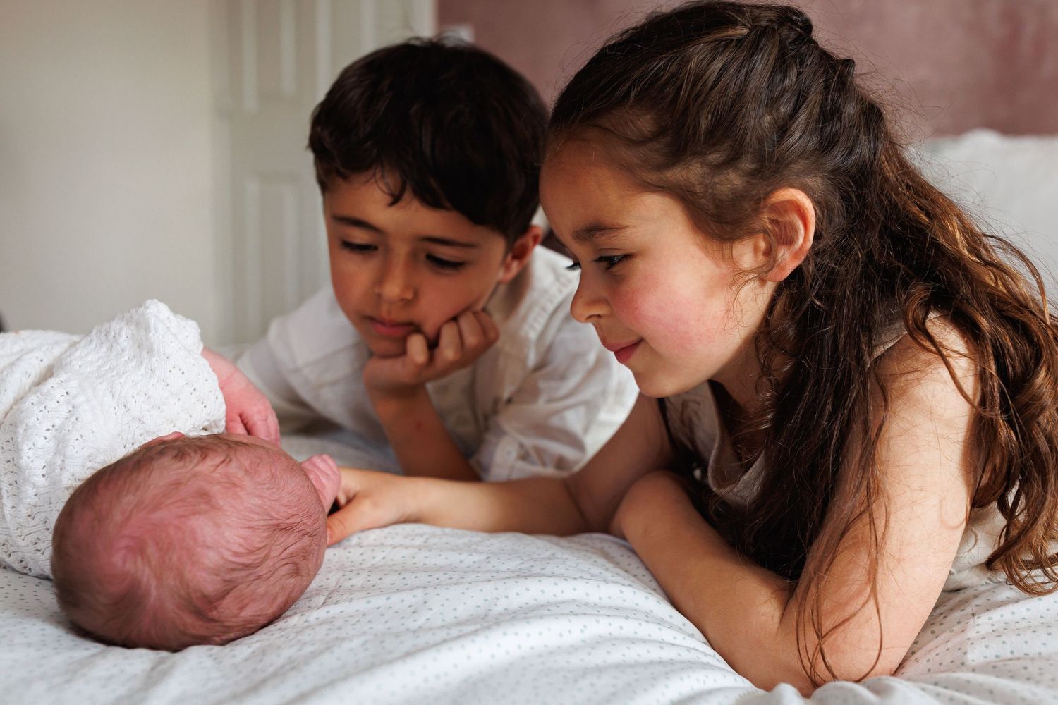 Two siblings lie on a bed, curiously and lovingly gazing at their newborn baby sibling.