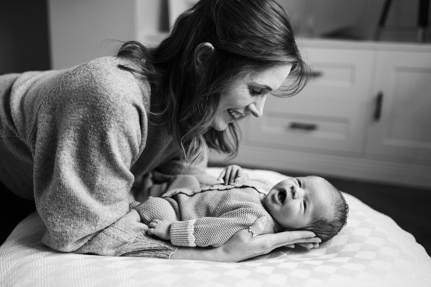 Black and white photo of a smiling mother leaning over her laughing newborn baby on a white bed.