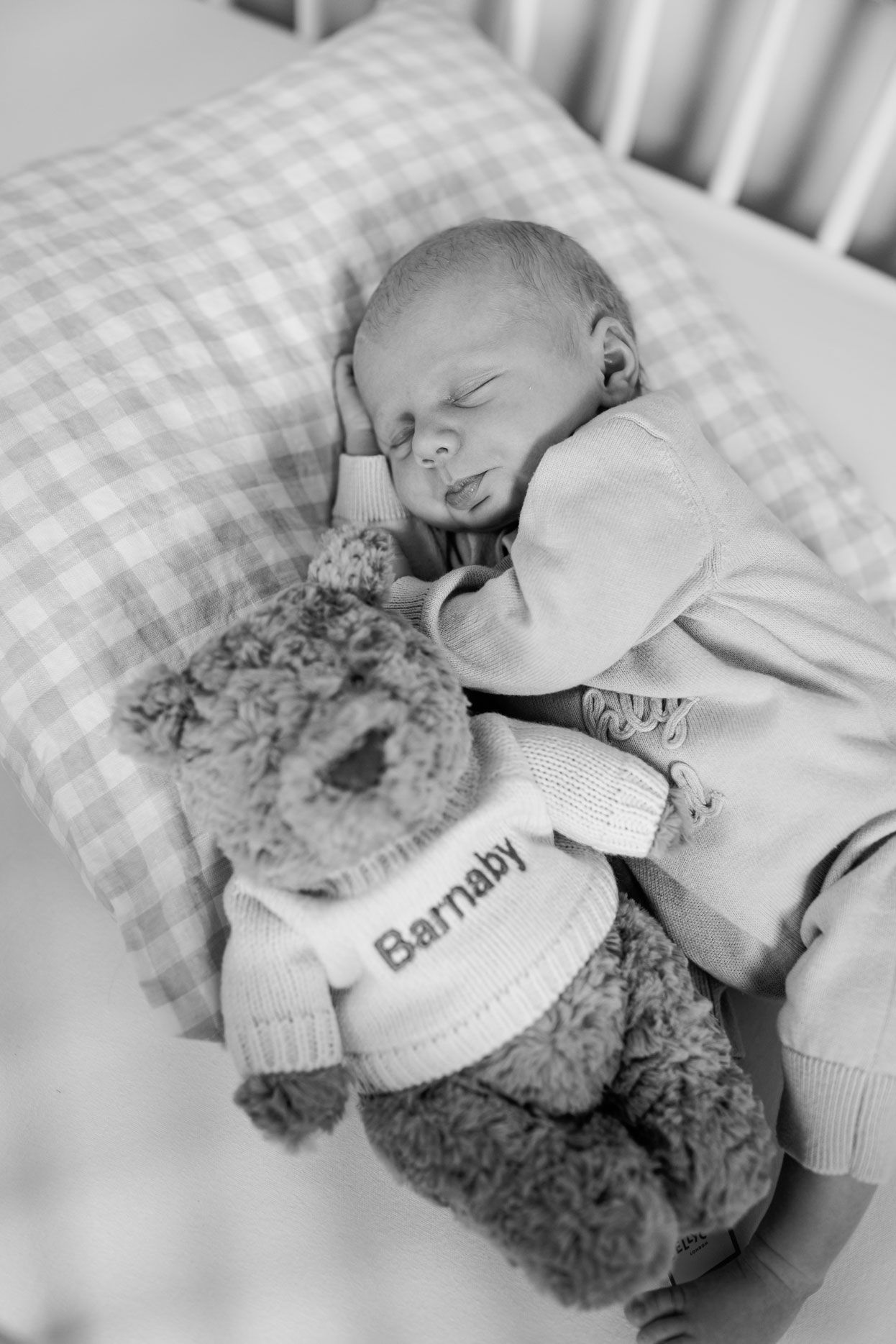 Black and white photo of a sleeping newborn baby on a checkered pillow in a crib, cuddling a teddy bear in a sweater that reads Barnaby.
