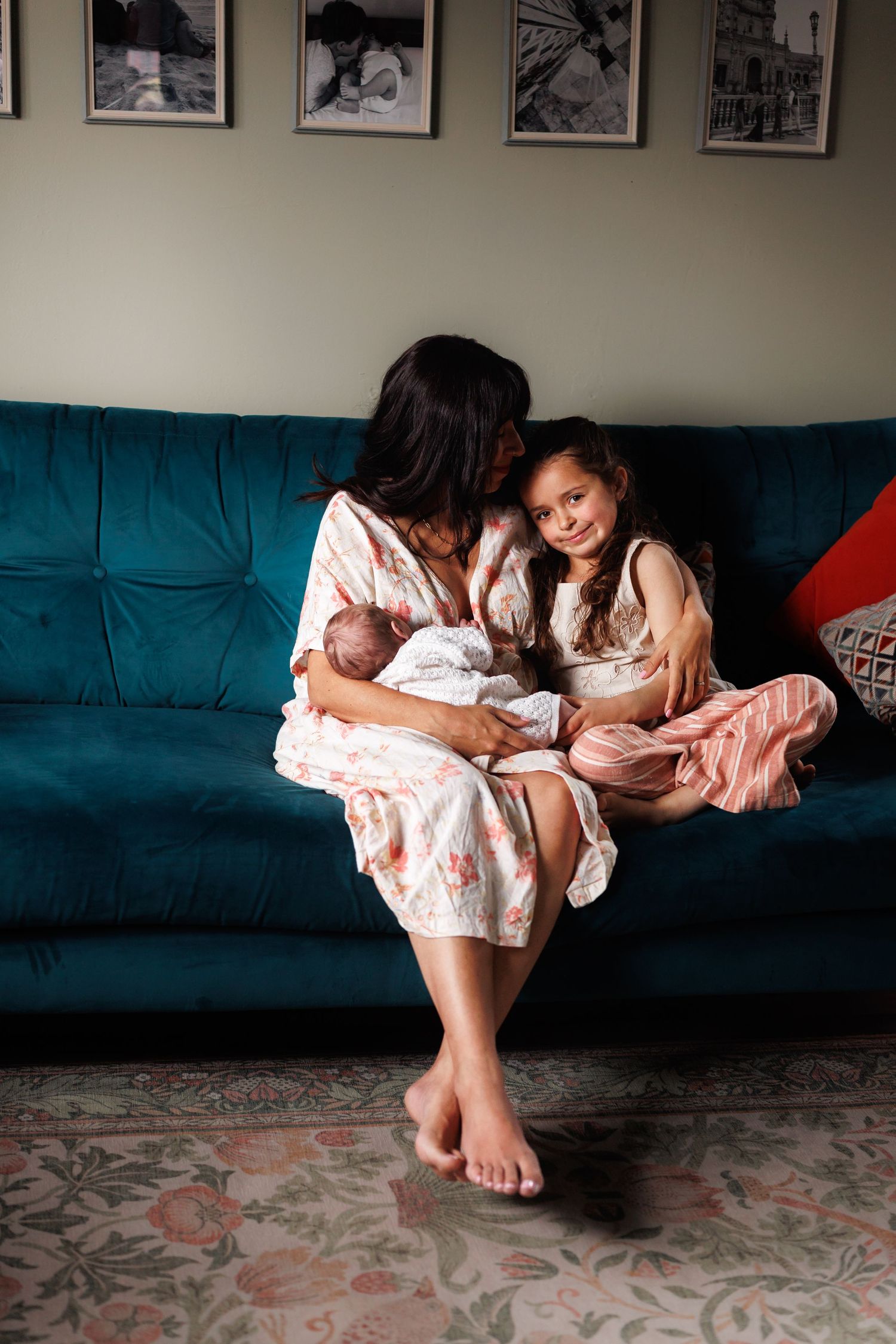 Mother in floral robe sitting on teal sofa cuddling newborn while young daughter smiles beside her.