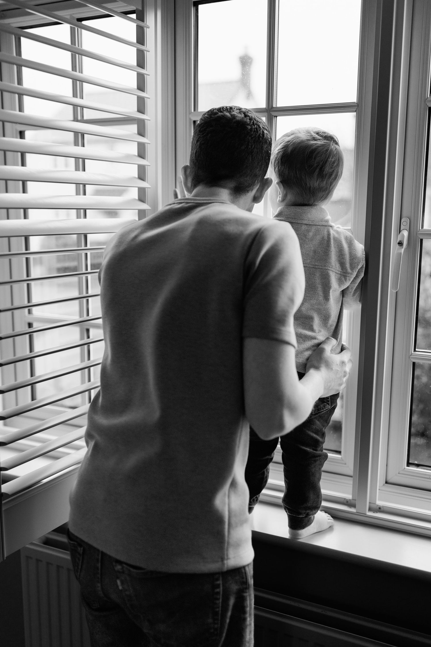 Black and white rear view of a father holding his toddler son while they look out a window together.