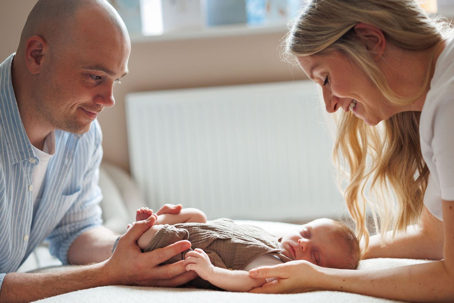 Two loving parents lean over their newborn baby lying on a soft surface in warm natural light.