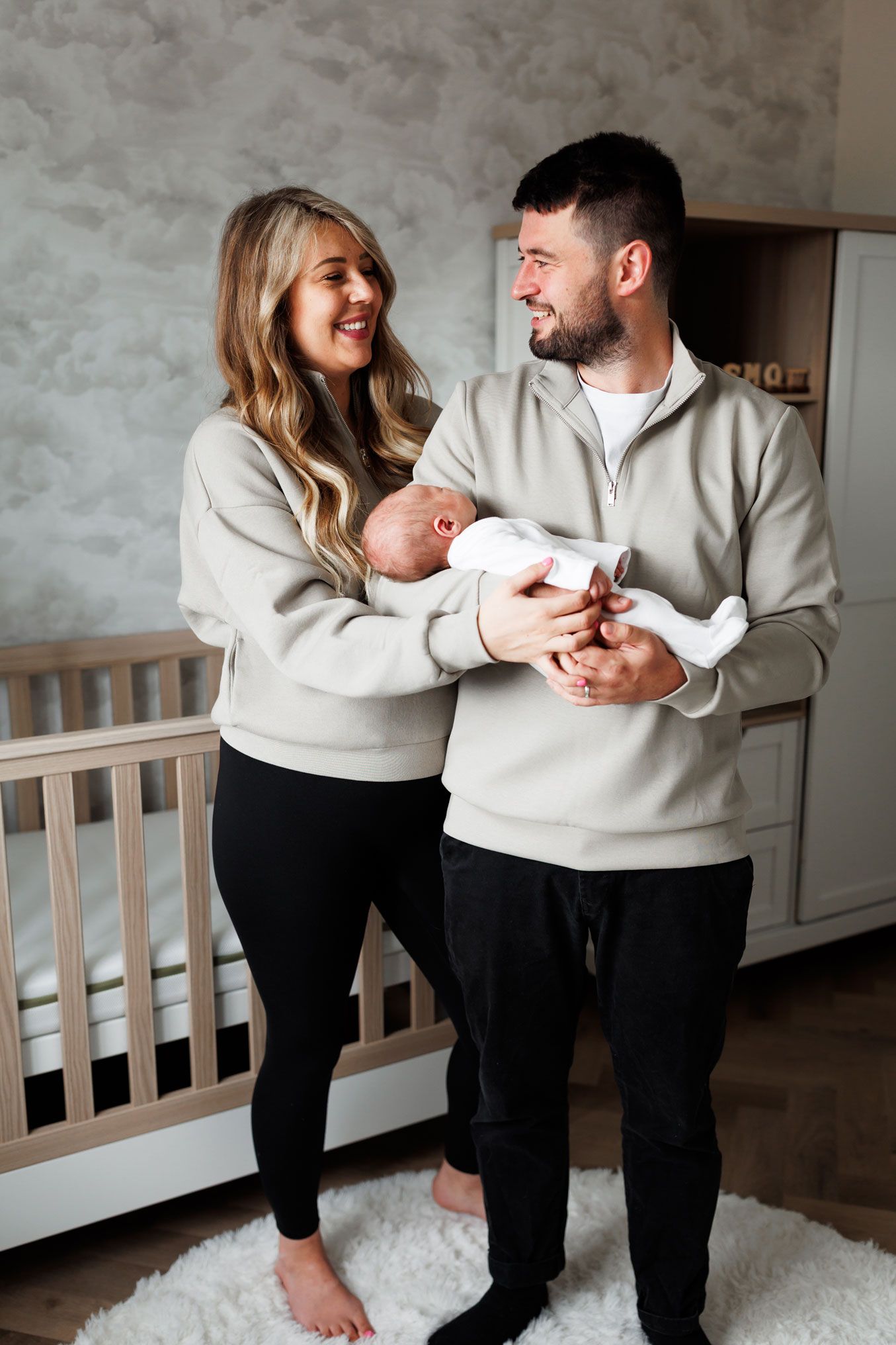 Smiling couple in matching grey sweatshirts and black pants holding their newborn baby in a nursery.