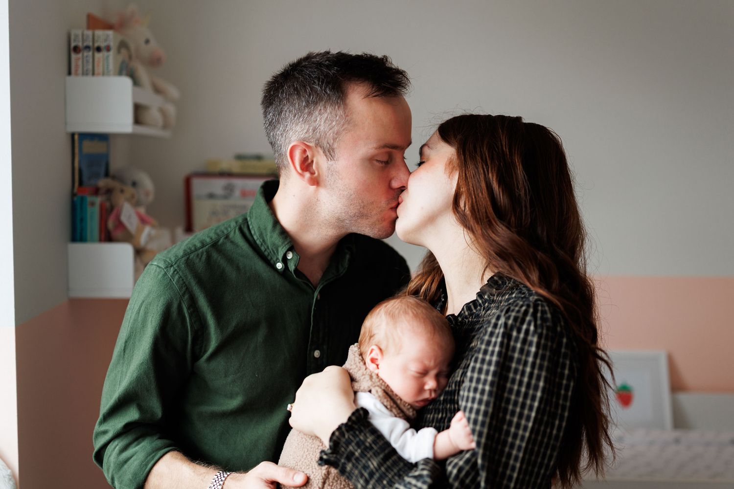 A man in a green shirt and a woman in a plaid top share a kiss while holding their sleeping newborn baby in a bright nursery.