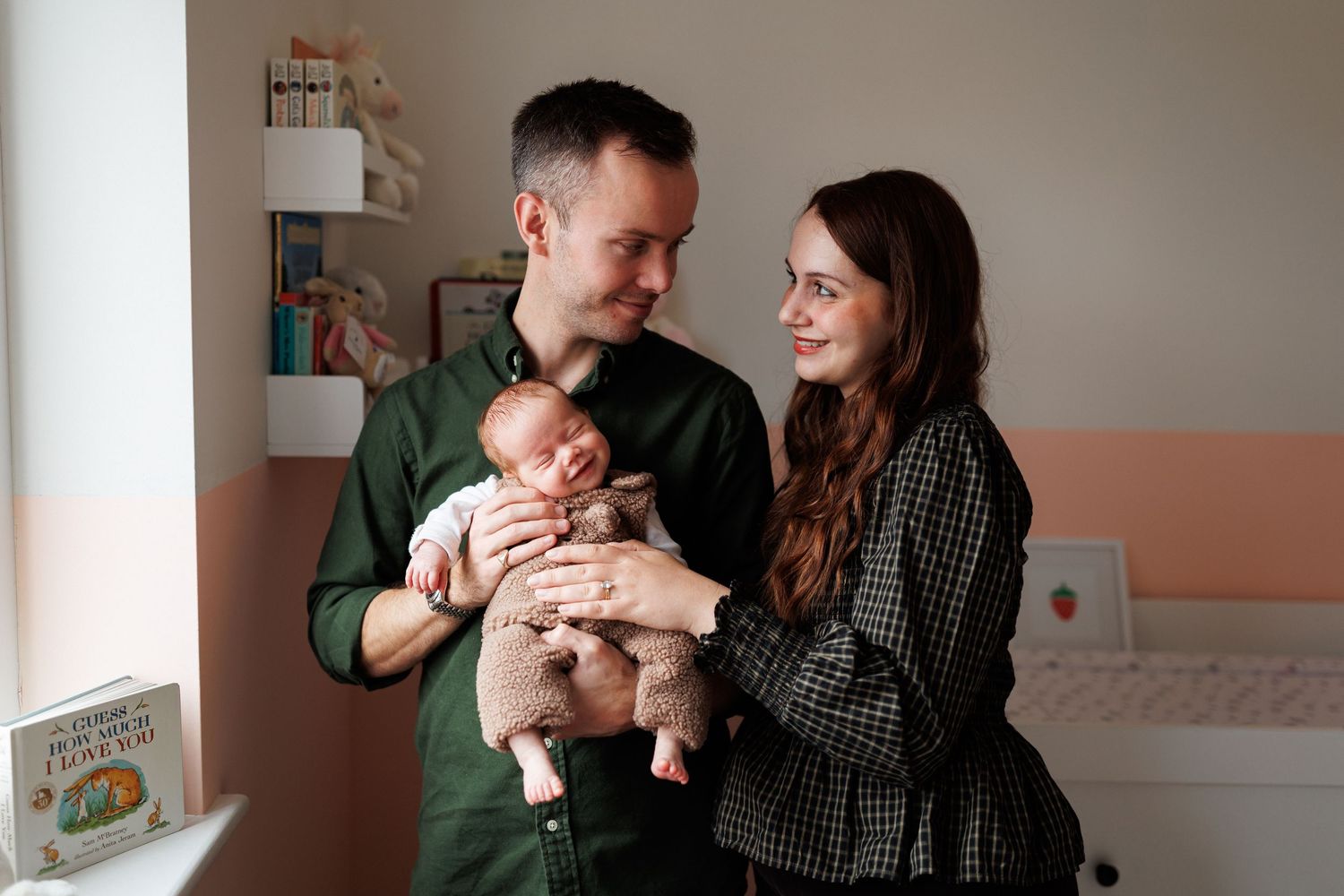 Smiling couple standing in a cozy nursery, gently holding their sleeping newborn baby in a cream onesie beside shelves and a white dresser.