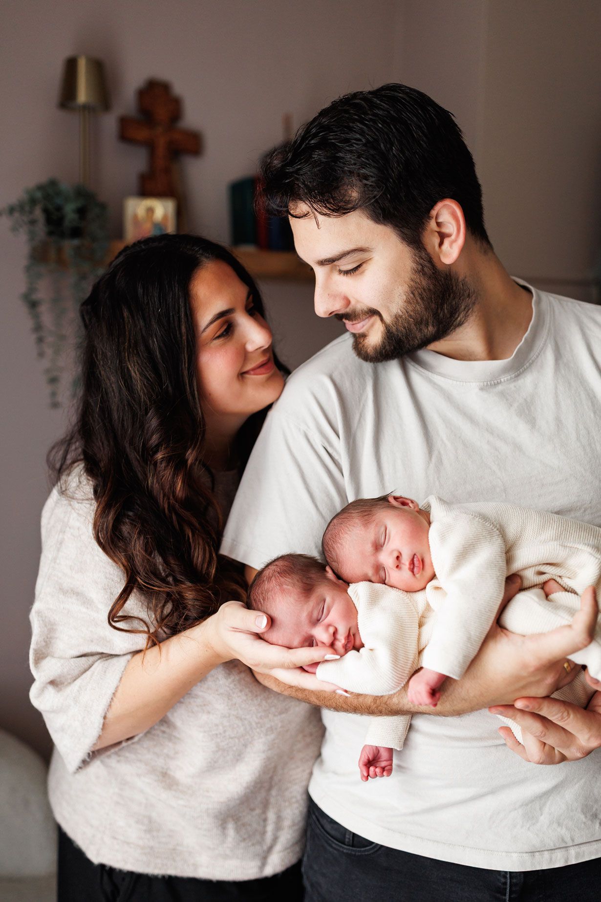 Smiling bearded man and woman with long dark hair gently hold two sleeping newborn twins near a wooden cross on a shelf.