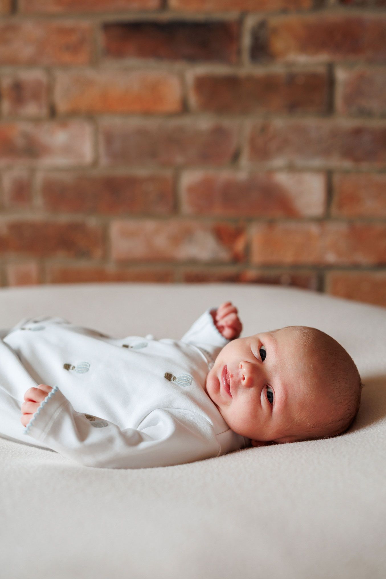 Newborn baby in a white embroidered outfit lying awake on a cream blanket in front of a rustic red brick wall.