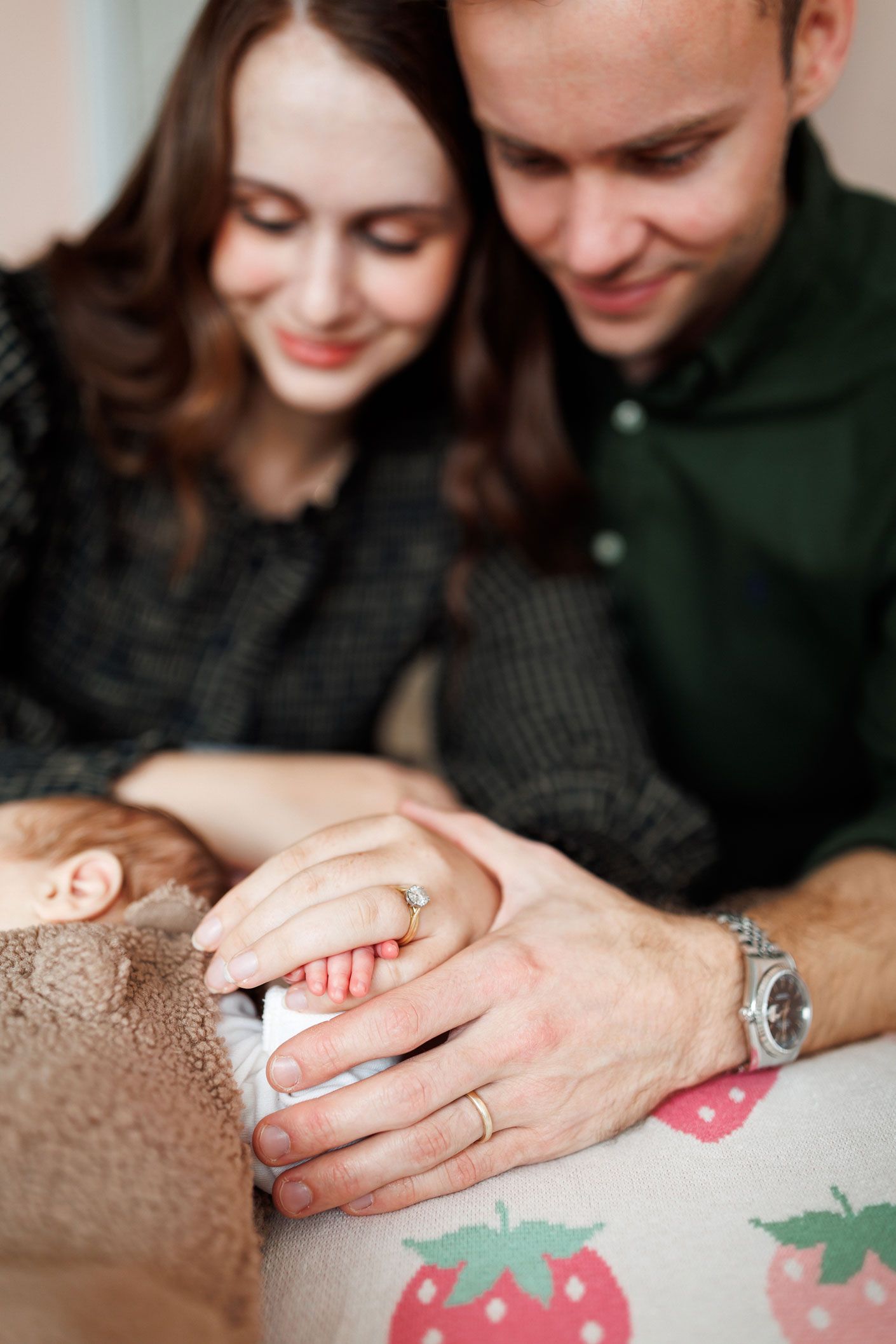 Smiling new parents gently hold their sleeping newborn baby's tiny hand, resting on a soft blanket with a strawberry pattern.