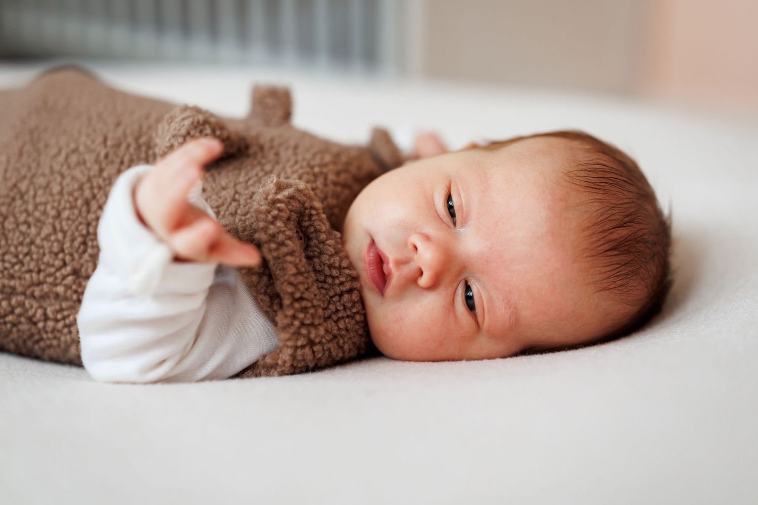 Newborn baby with light reddish hair wearing a soft brown fleece sleeper, lying awake on a white sheet with one arm stretched out.