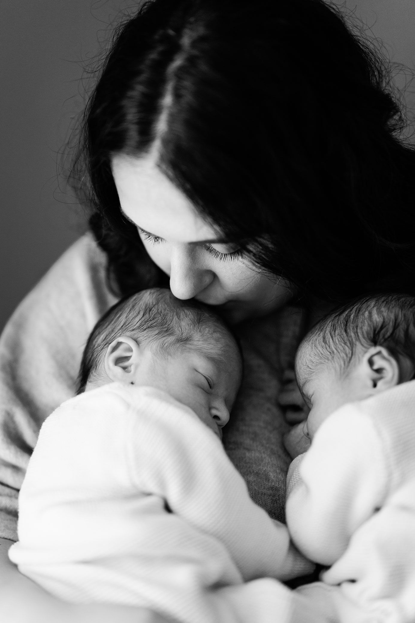 Black and white portrait of a mother tenderly kissing the forehead of one of her two sleeping newborn twins cradled in her arms.
