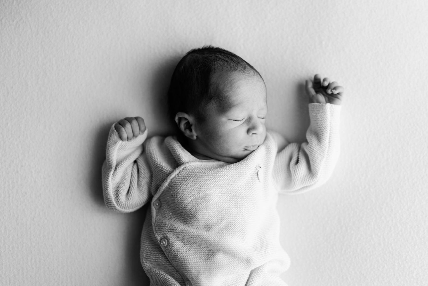 Black and white photo of a sleeping newborn baby in a soft wrap top, lying on their back with tiny fists raised on a plain backdrop.