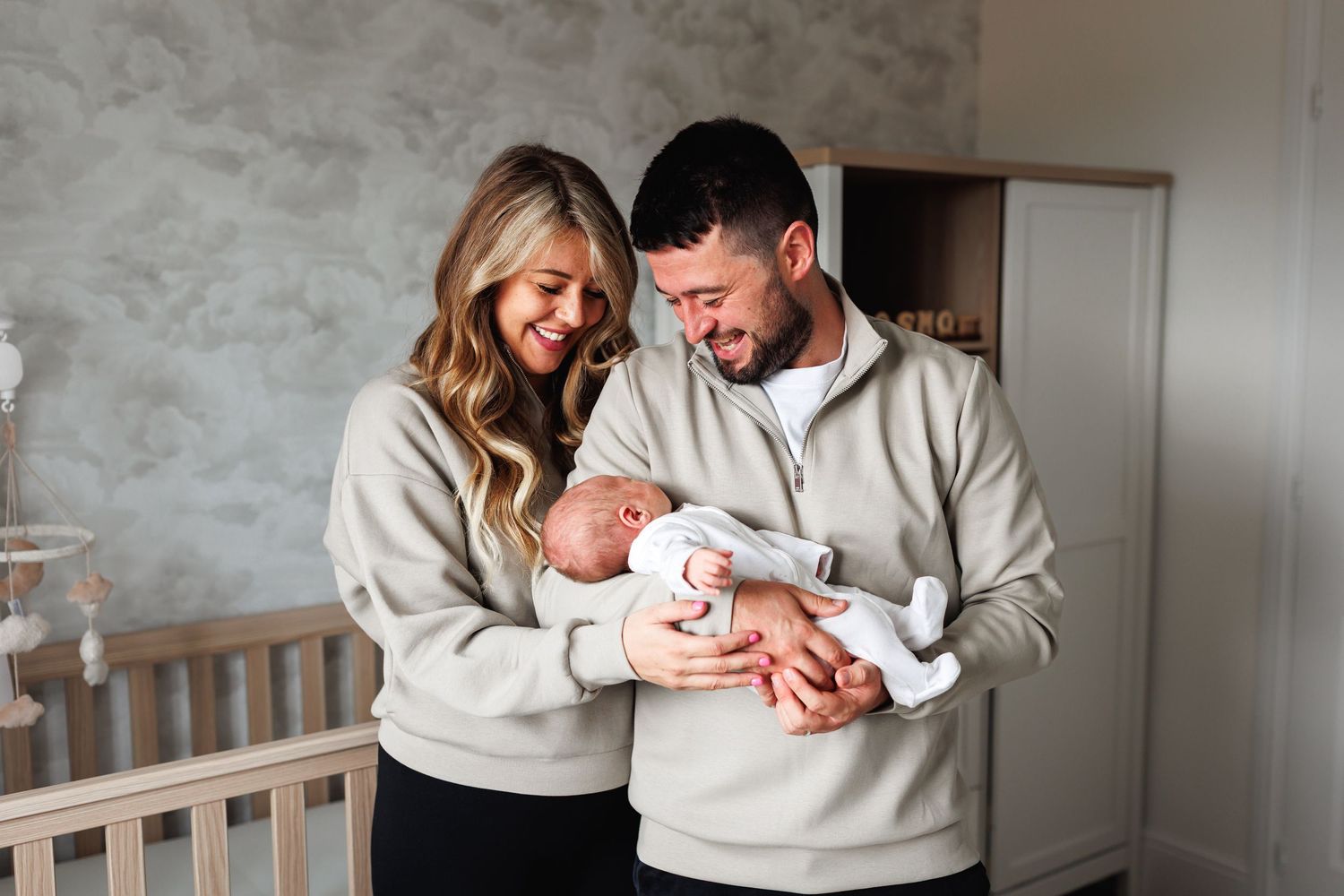 Matching couple in beige hoodies smiling at their newborn baby in a nursery room.