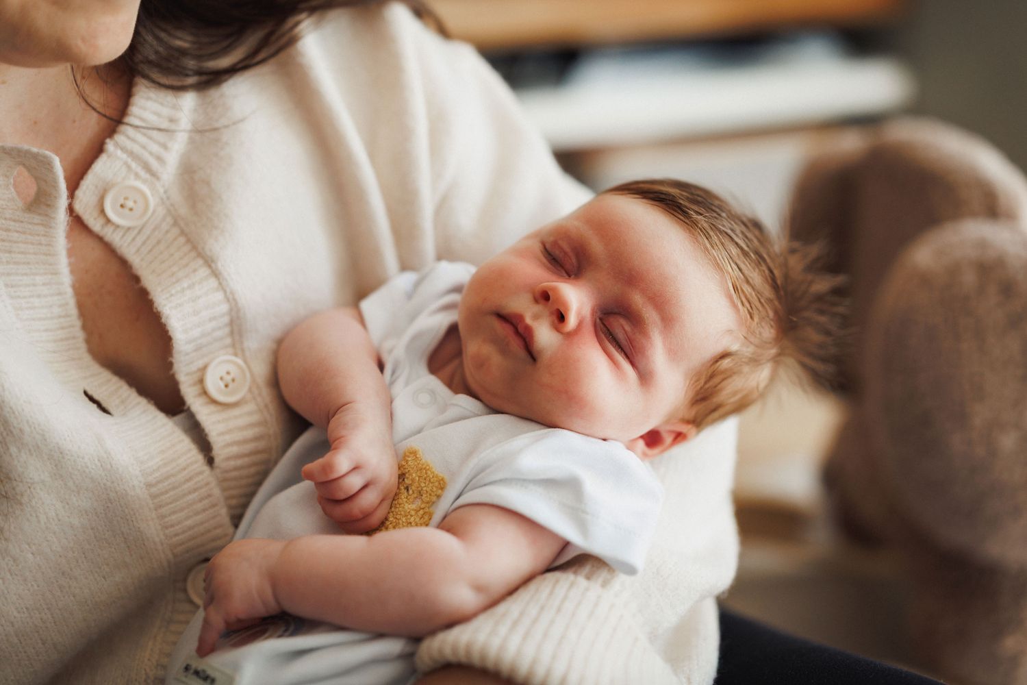 Newborn baby sleeping peacefully in mother's arms, dressed in white onesie, soft natural light.