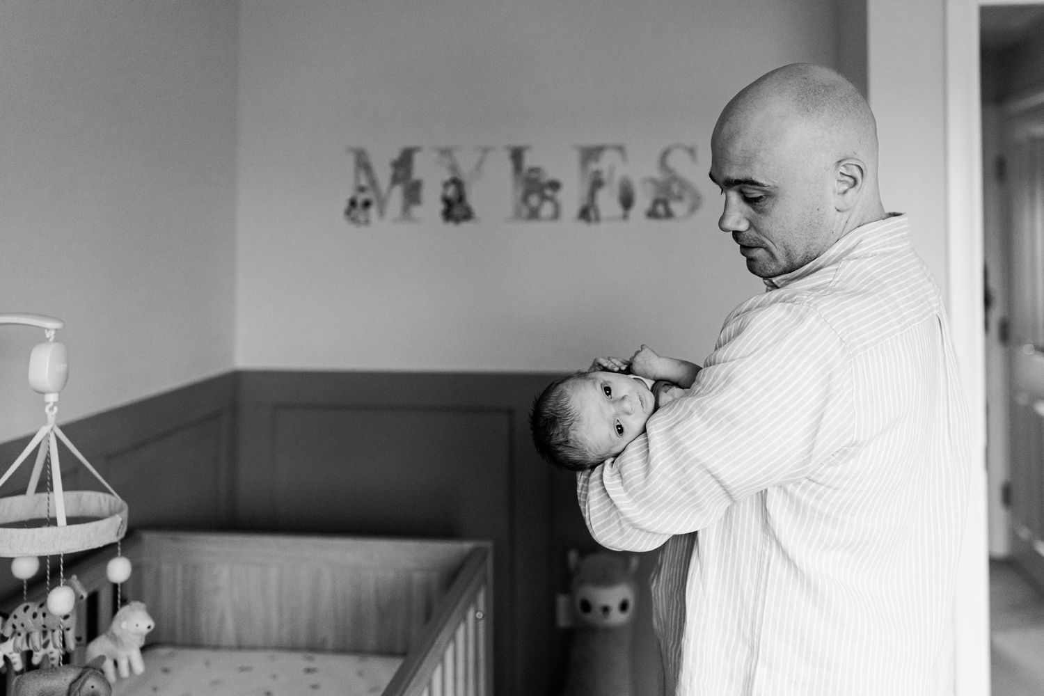 Bald father tenderly holds newborn baby in nursery with name MYLES on wall, black and white photo.