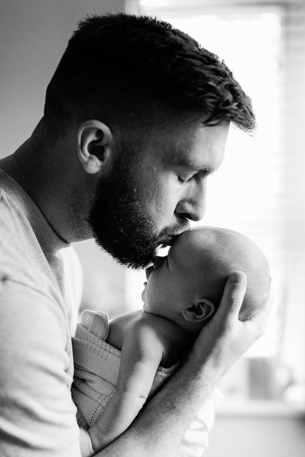 Black and white photo of a bearded father tenderly kissing his newborn baby on the forehead.