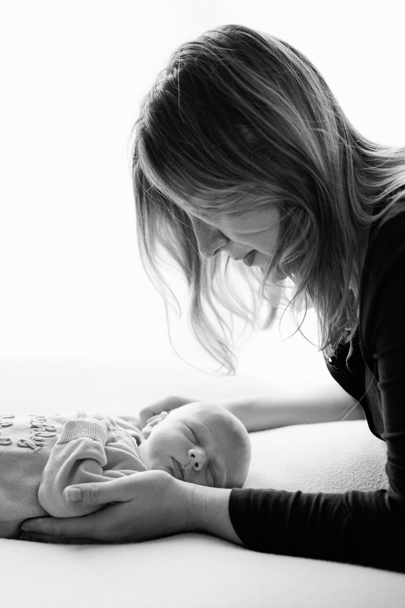 Black and white portrait of a mother with long hair leaning over her sleeping newborn baby while gently holding the infant's hands.