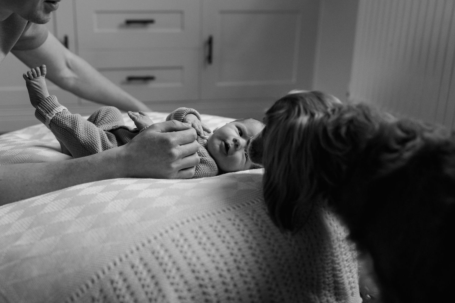 Black and white photo of parents and a dog gathered around a newborn baby lying on a white bed.