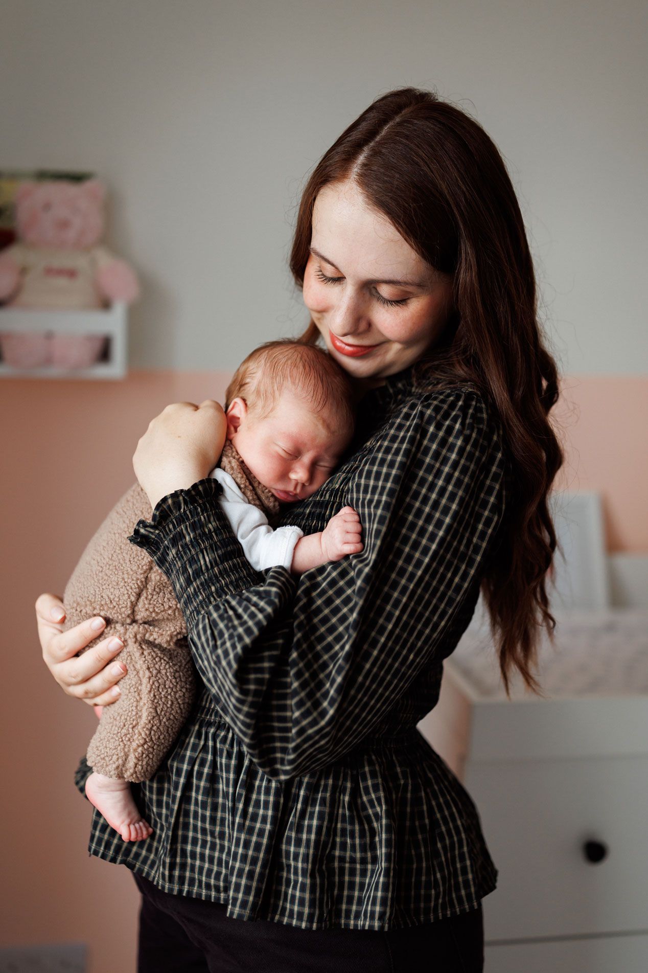 A smiling mother in a black plaid blouse gently cradles her sleeping newborn baby in a cozy nursery with soft peach walls.